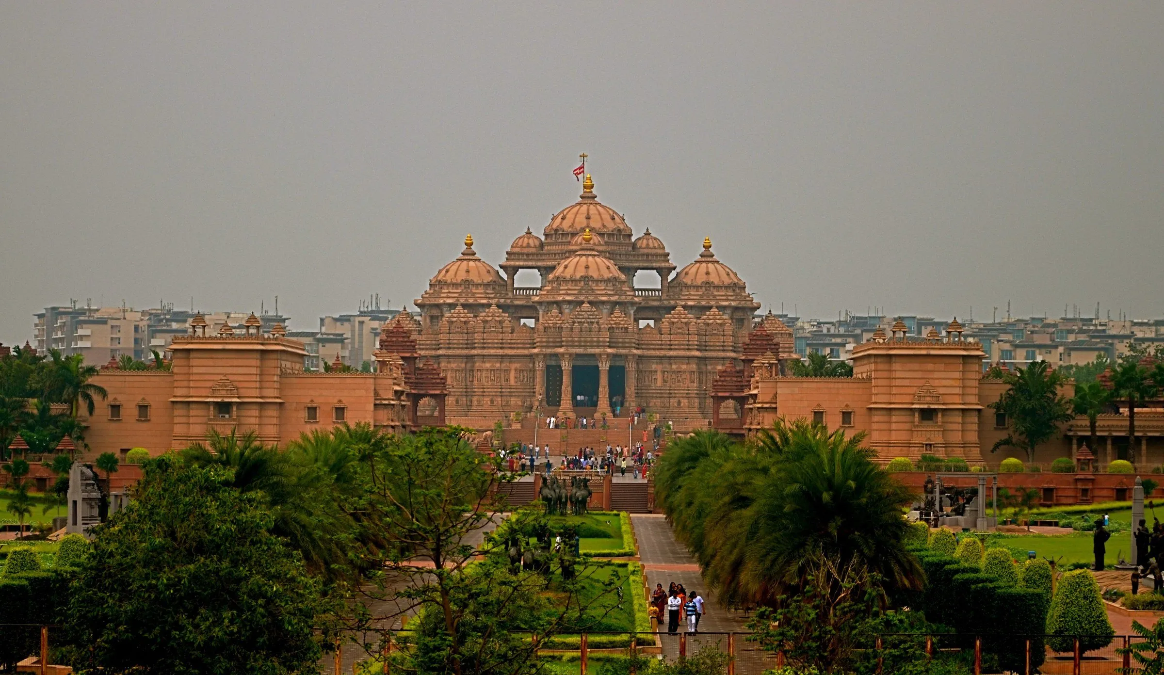 Fachada de arenisca rosa del templo Akshardham con cúpulas y pináculos, vista desde un jardín con palmeras y visitantes.