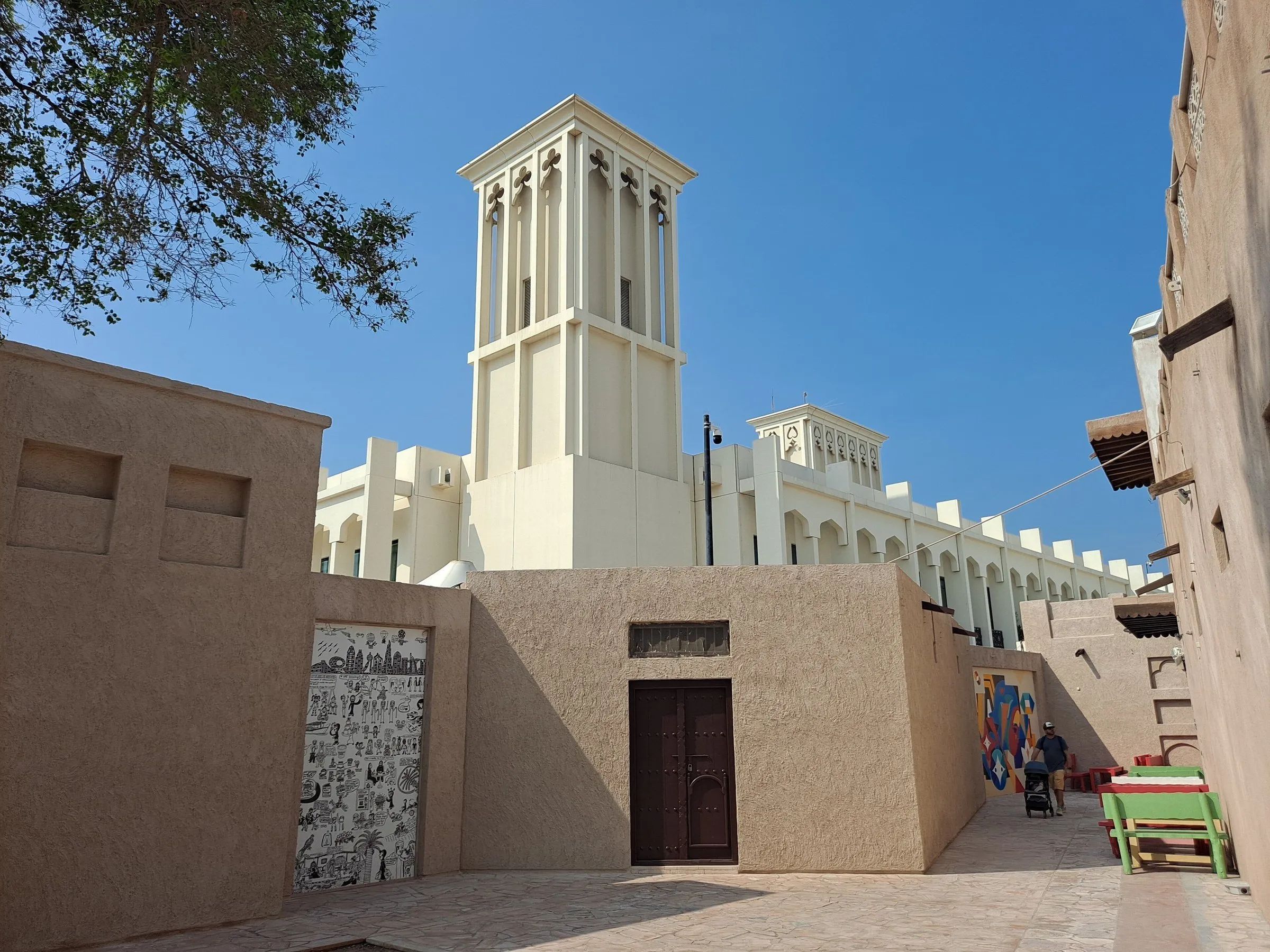Al Fahidi lane between sand-colored mud walls, with a white wind tower and blue sky in the background.