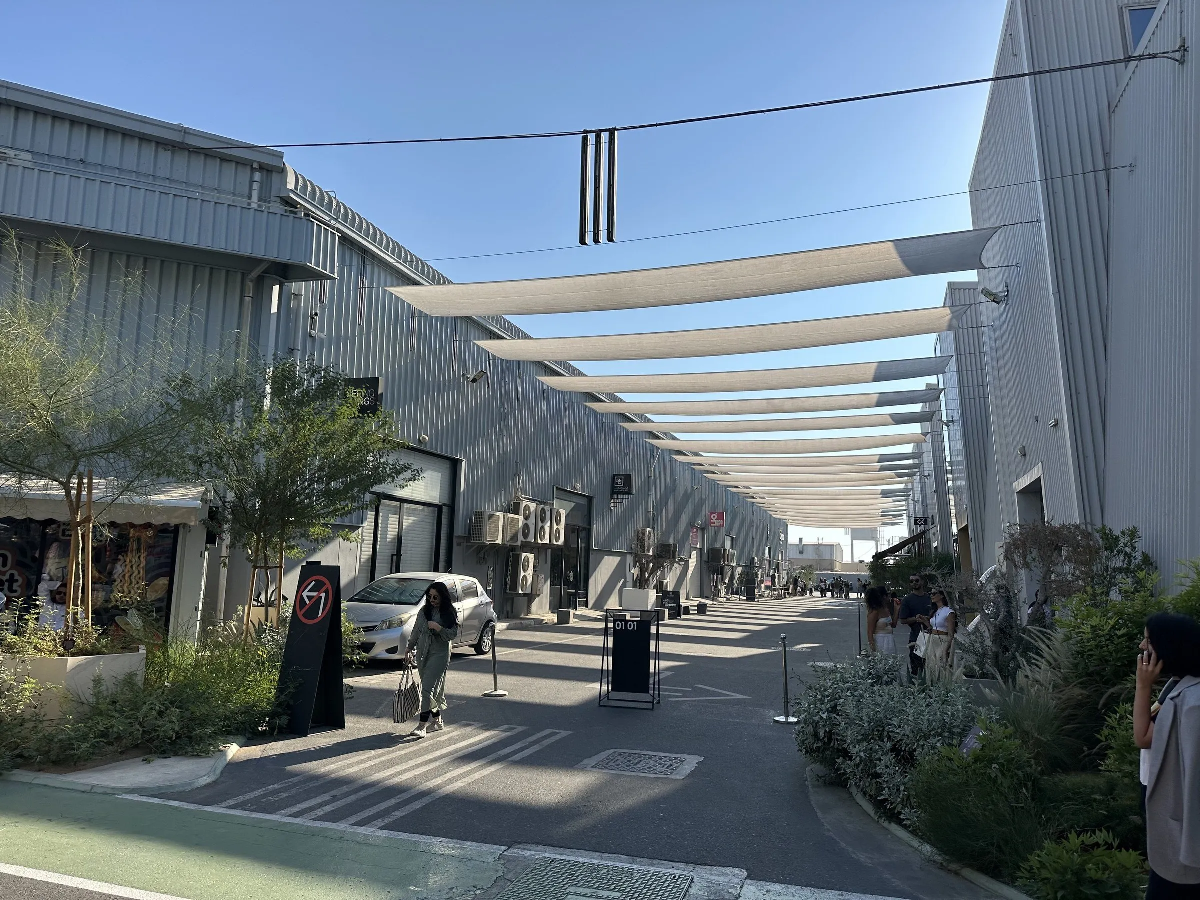 Street between metal warehouses at Alserkal Avenue in Dubai, covered by white shade sails, with plants and few people.