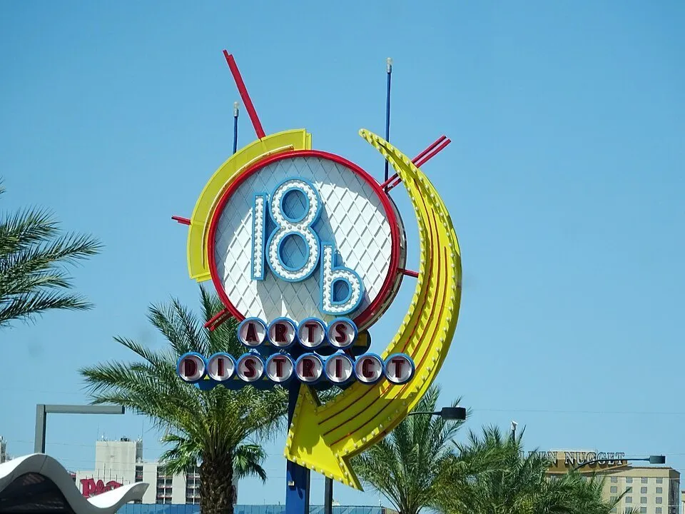 Yellow, red and blue 18b Arts District sign with palm trees and a blue sky behind.