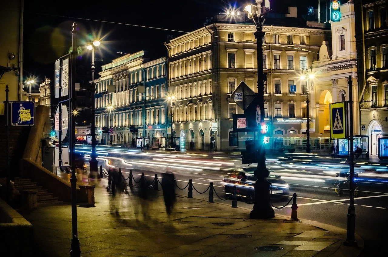 Avenida Nevsky à noite, com fachadas iluminadas, postes e rastros dos veículos junto à calçada.