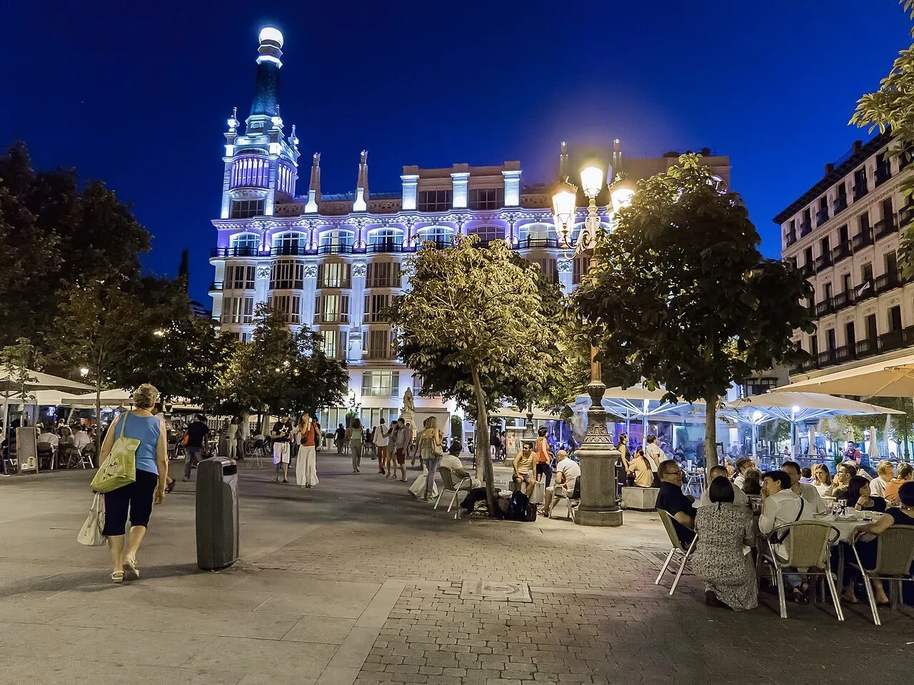 Plaza de Santa Ana at night, with a lit building, trees, restaurant tables and pedestrians.