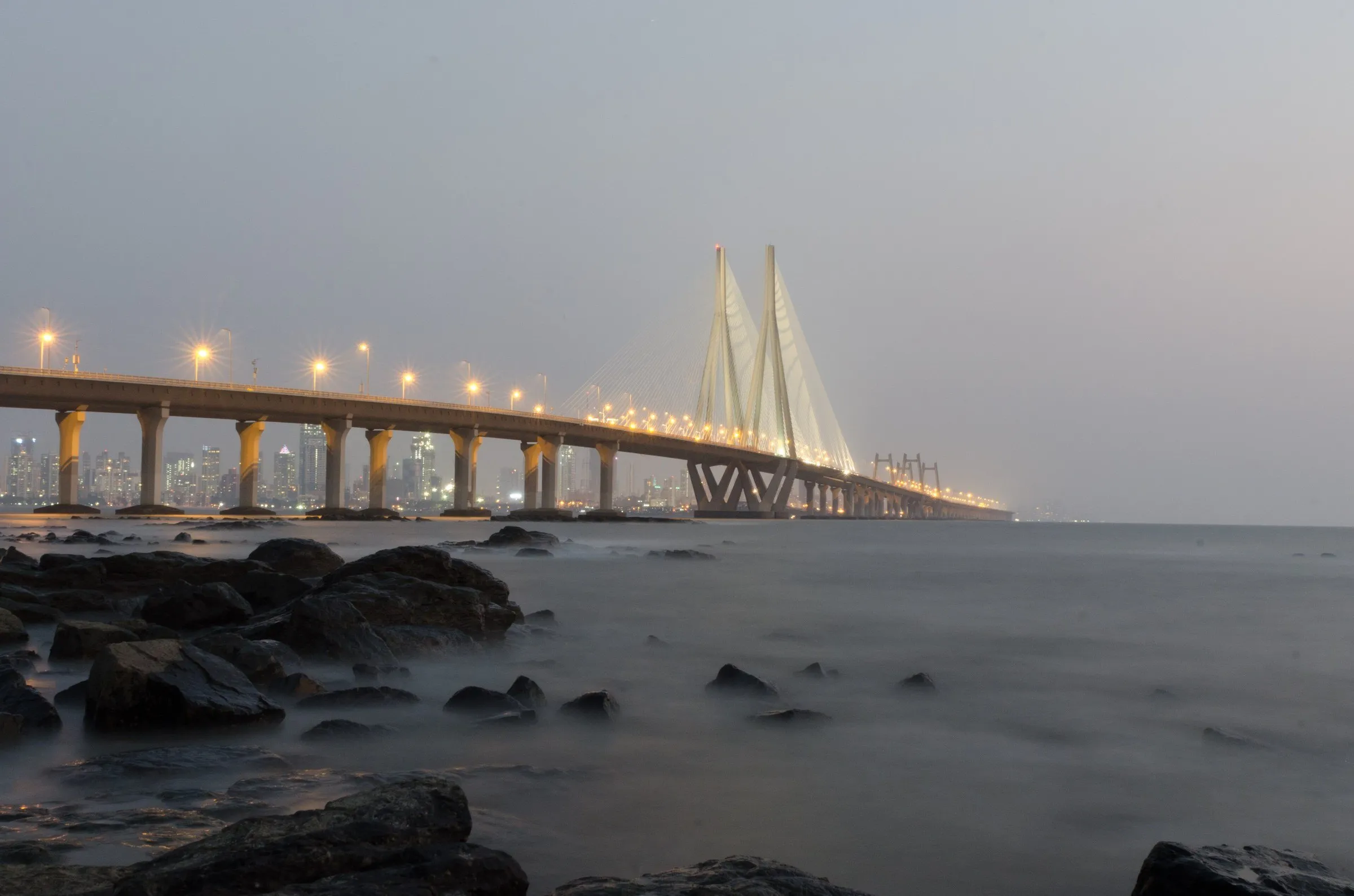 Ponte Bandra-Worli Sea Link iluminada ao entardecer, vista das rochas de Bandra, em Mumbai, com o mar em longa exposição.
