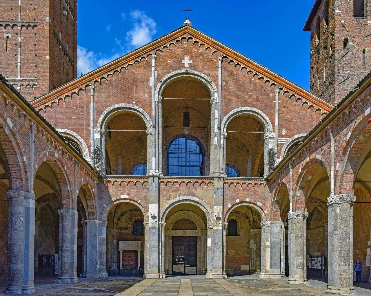 Brick façade of the Basilica of Sant’Ambrogio seen from the courtyard between arcades, under a blue sky.
