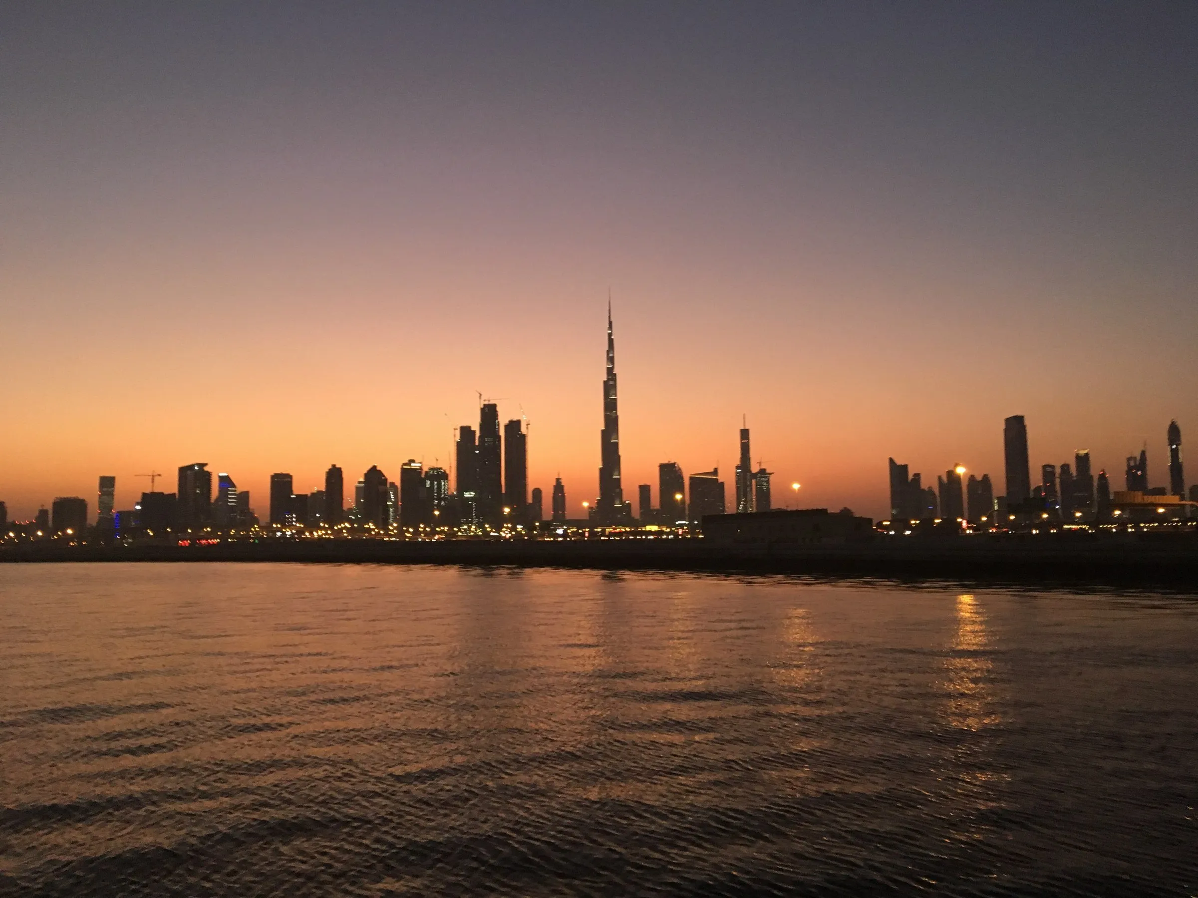 Dubai skyline with the Burj Khalifa silhouette over the sea, under an orange and purple sky just after sunset.