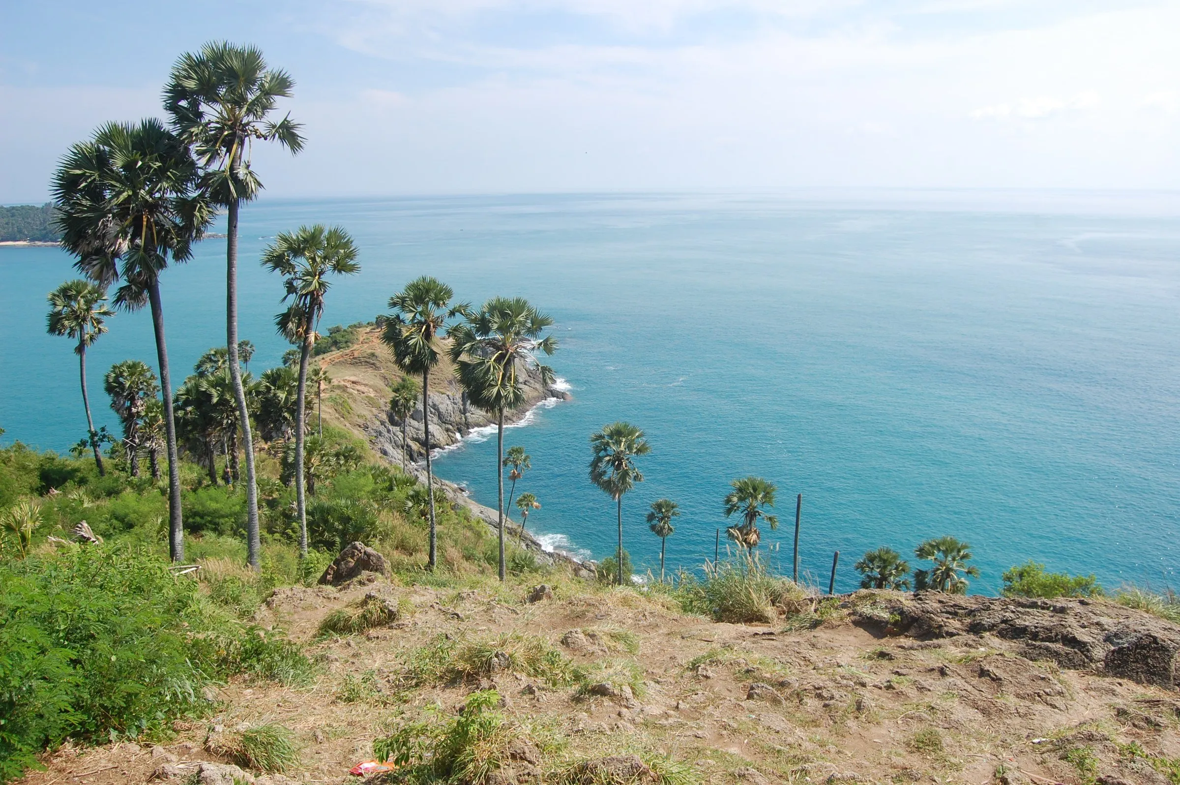 Cabo Promthep, em Phuket, visto do alto, com palmeiras, ponta de terra avançando no mar turquesa e ilhota ao fundo.