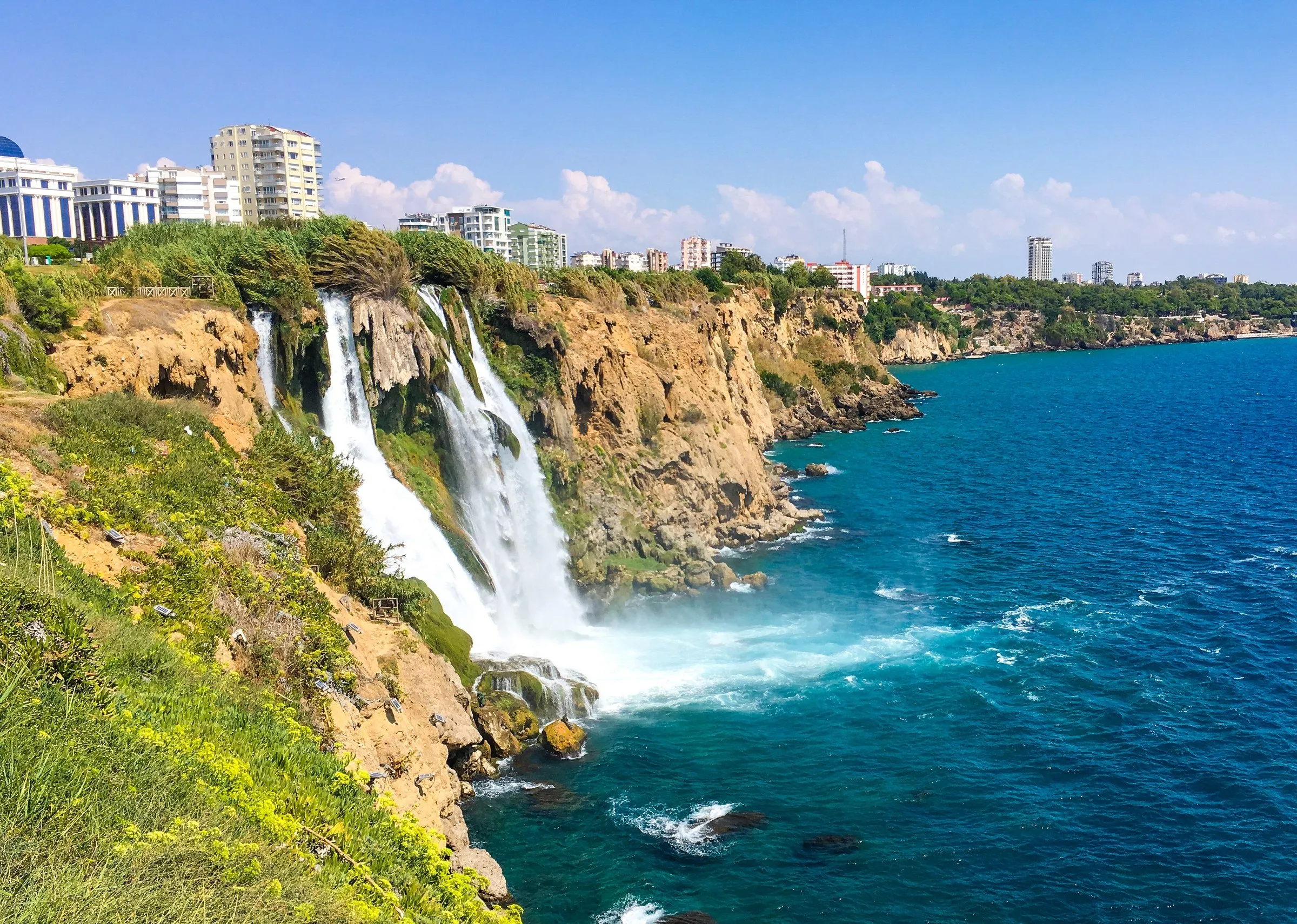 Cachoeira Düden de Baixo despencando de um penhasco coberto de mato no mar azul, com prédios da cidade no alto à esquerda.