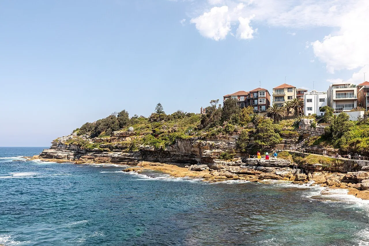 Stretch of the Bondi to Bronte walk beside rocks and blue sea, with a railing and houses on the hillside.