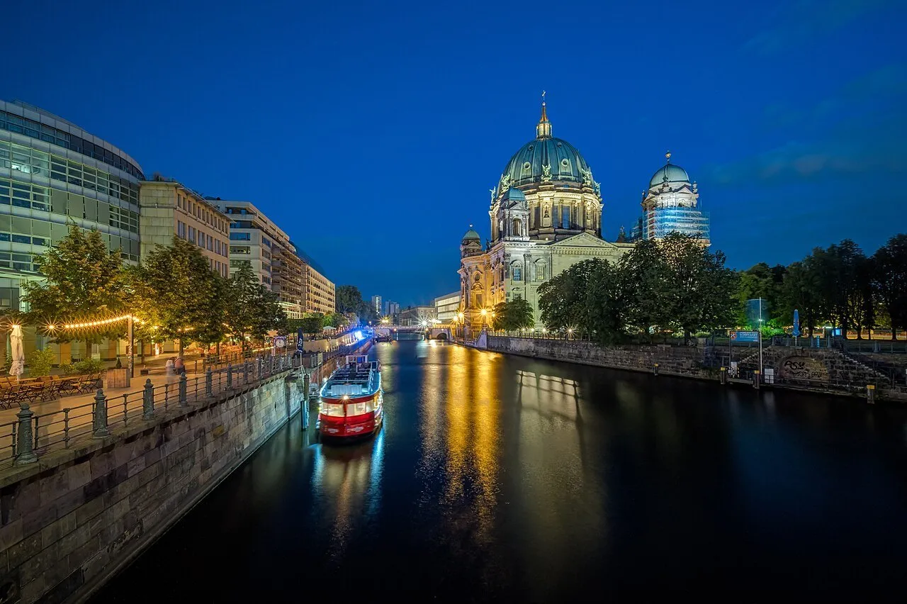 Illuminated Berlin Cathedral by the Spree, with a red boat and golden reflections in the water.