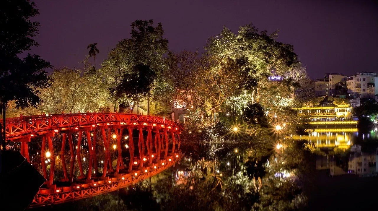 The red The Huc Bridge lit up at night, with trees and reflections in Hoan Kiem Lake.