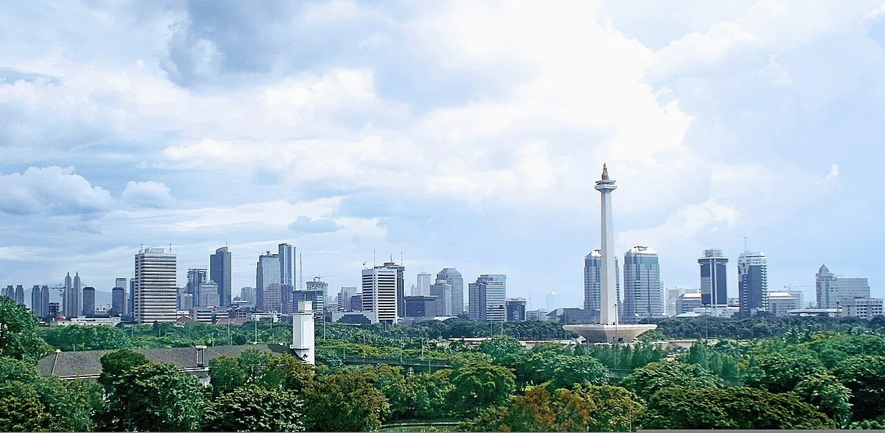 Panorama of Monas among trees, with Jakarta buildings behind and a cloudy sky.