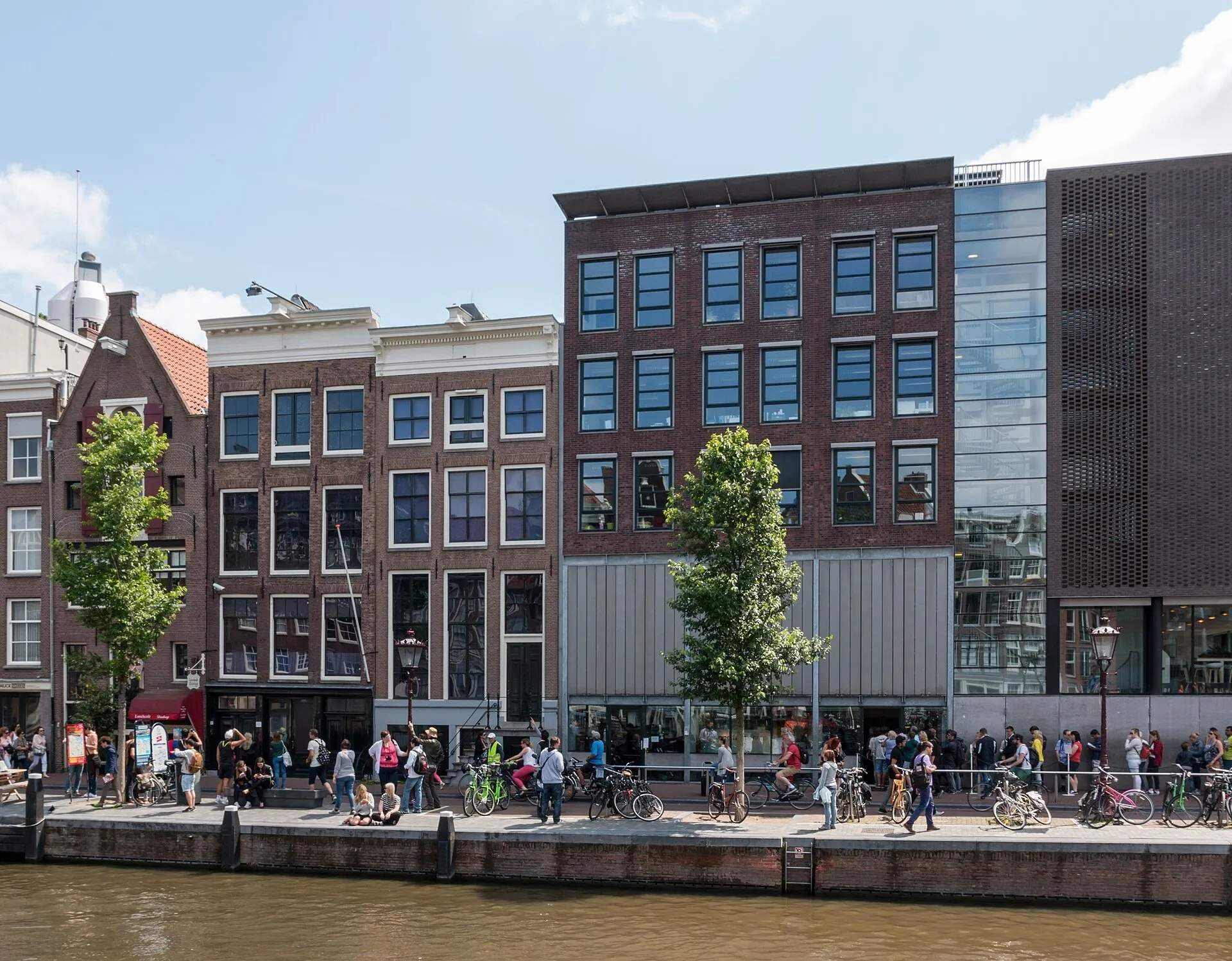 Fachada histórica da Casa de Anne Frank e ampliação do museu, com canal e pedestres à frente.