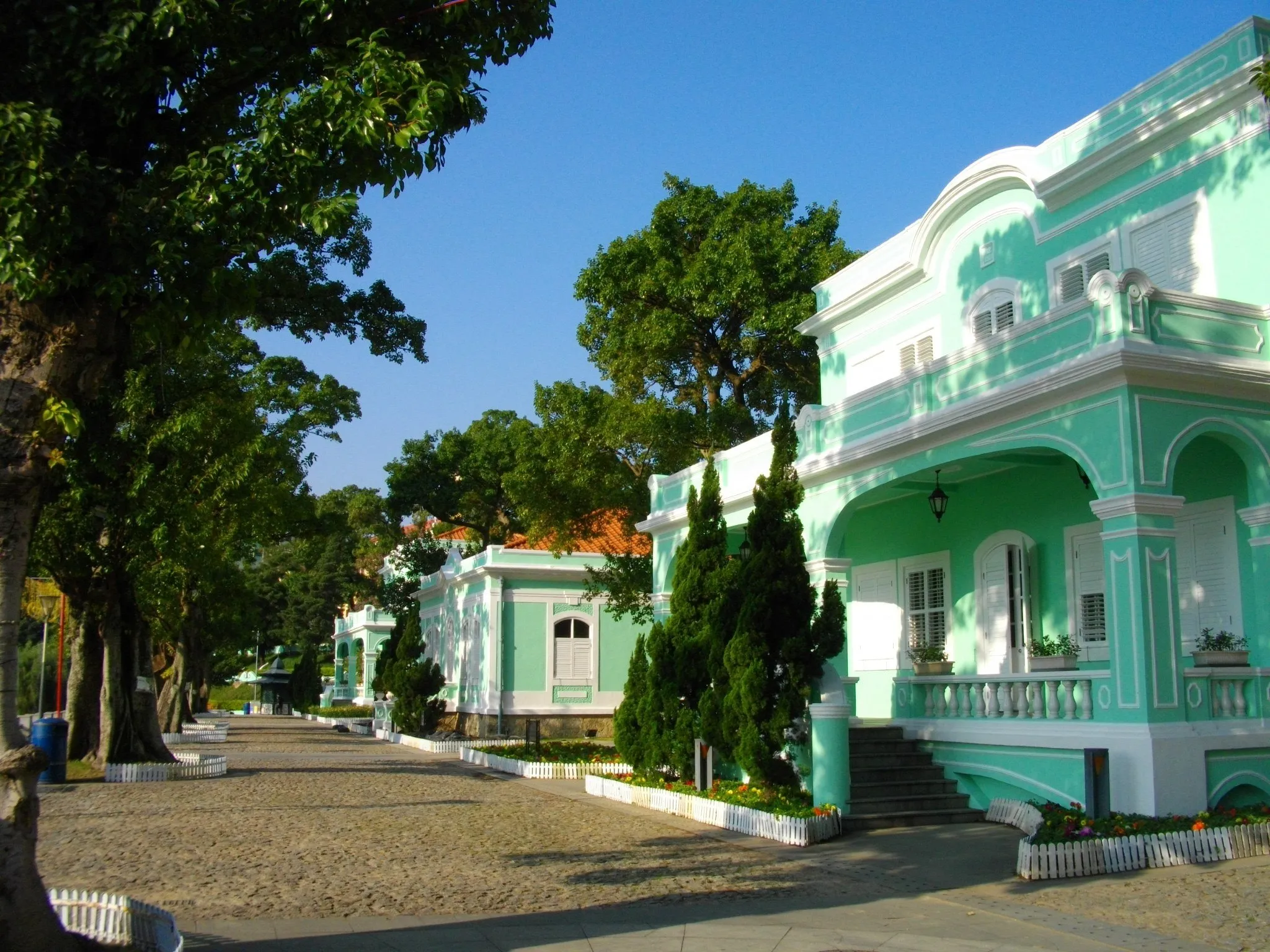 Casa-museo verde agua de dos plantas con balcón, junto a un camino de piedra bajo árboles y cielo azul.