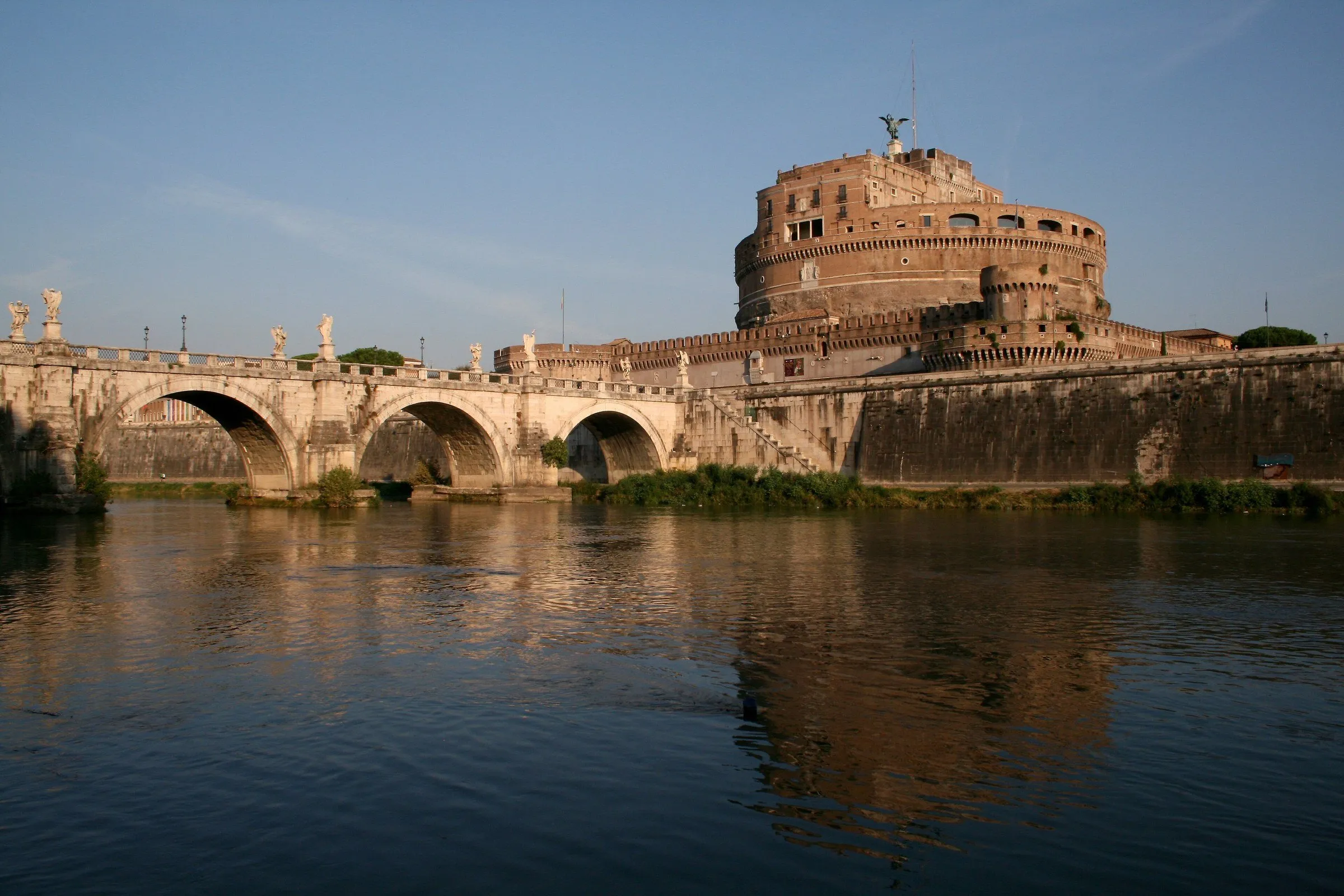 Castel Sant'Angelo and the stone arch bridge over the Tiber river, reflected in the water in golden light.