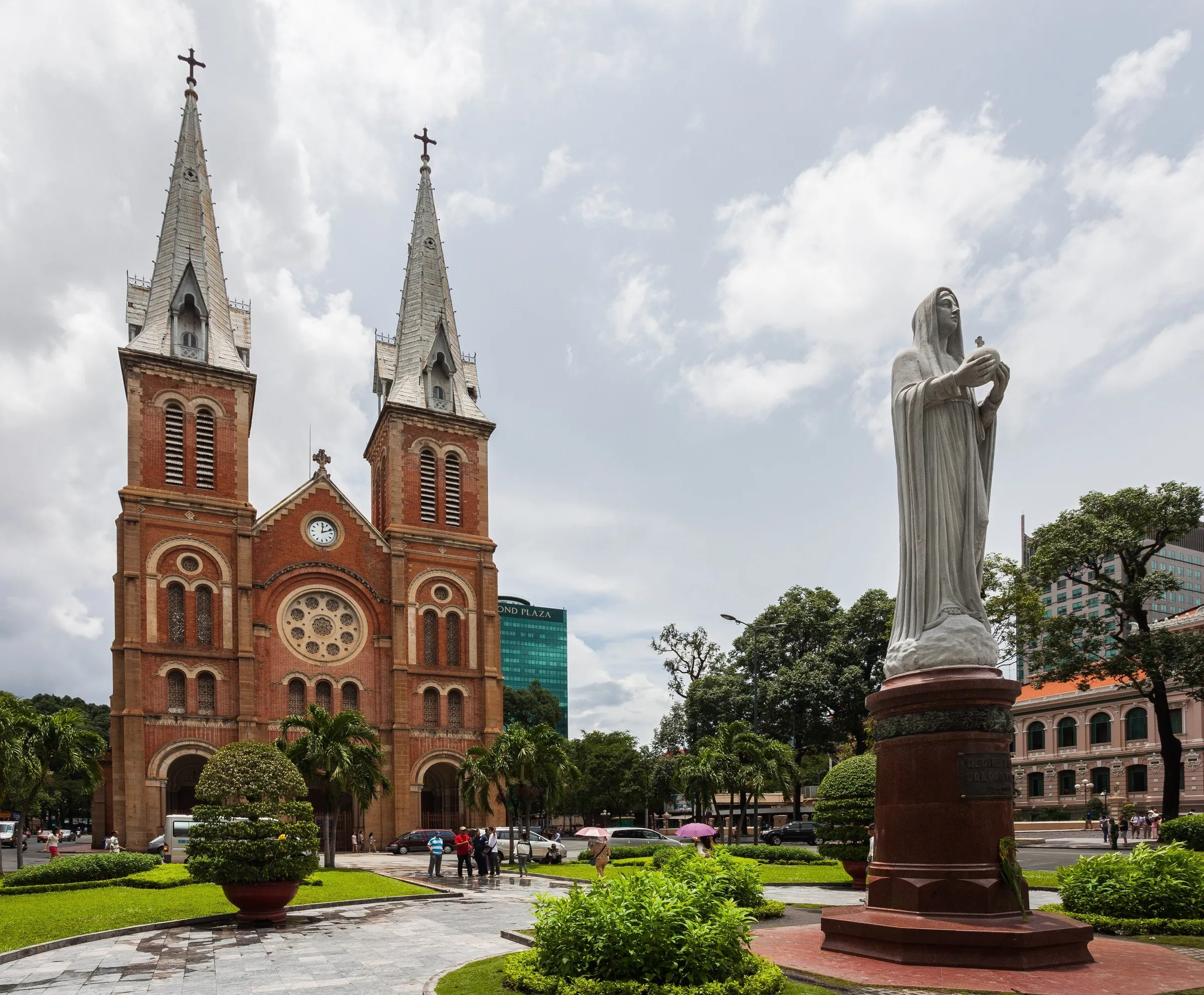 Fachada de tijolos da Catedral de Notre-Dame de Saigon com duas torres e estátua no jardim à direita.