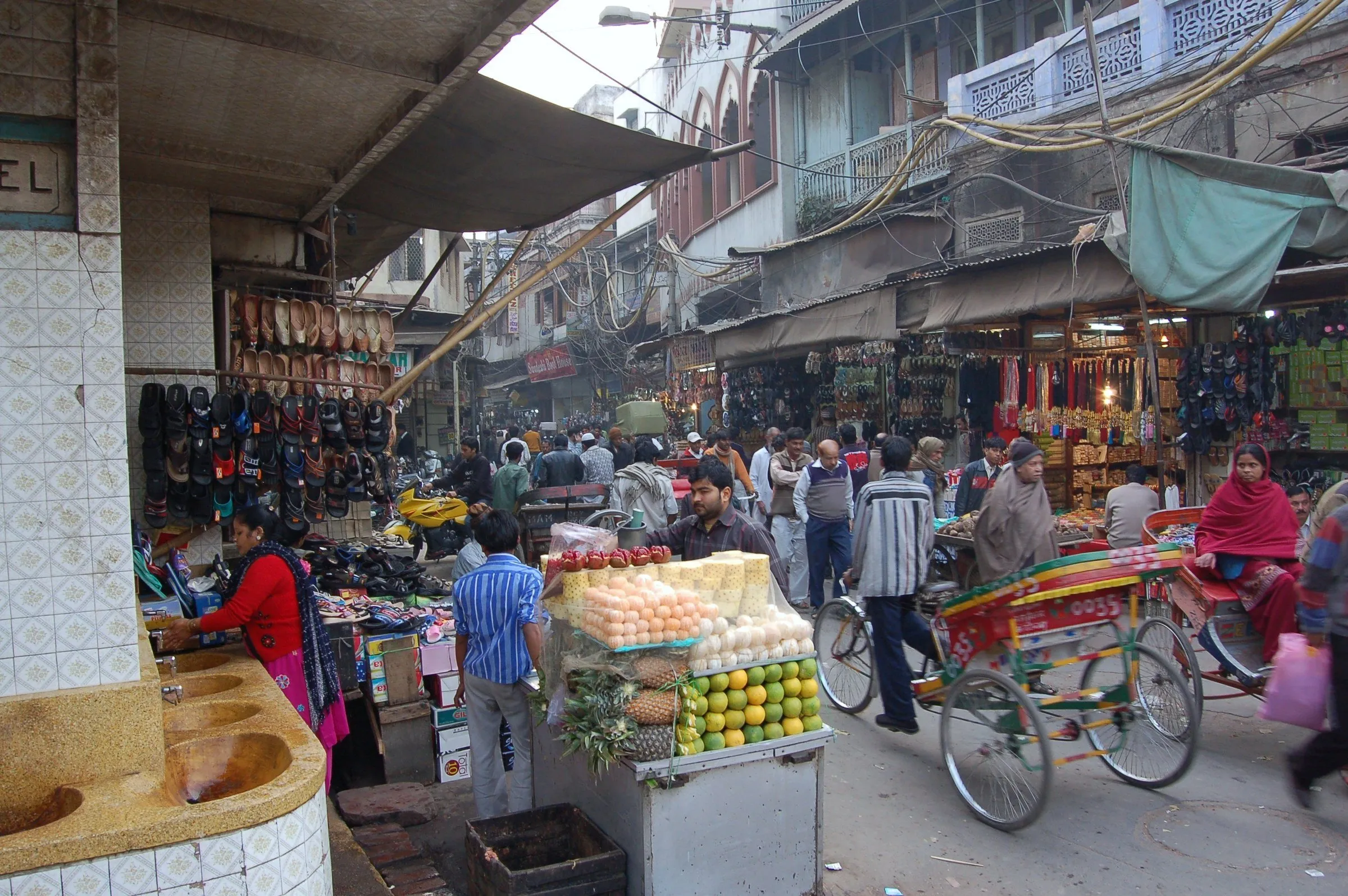 Calle concurrida del mercado de Chandni Chowk, con puestos de zapatos y fruta, un rickshaw colorido y gente bajo toldos.