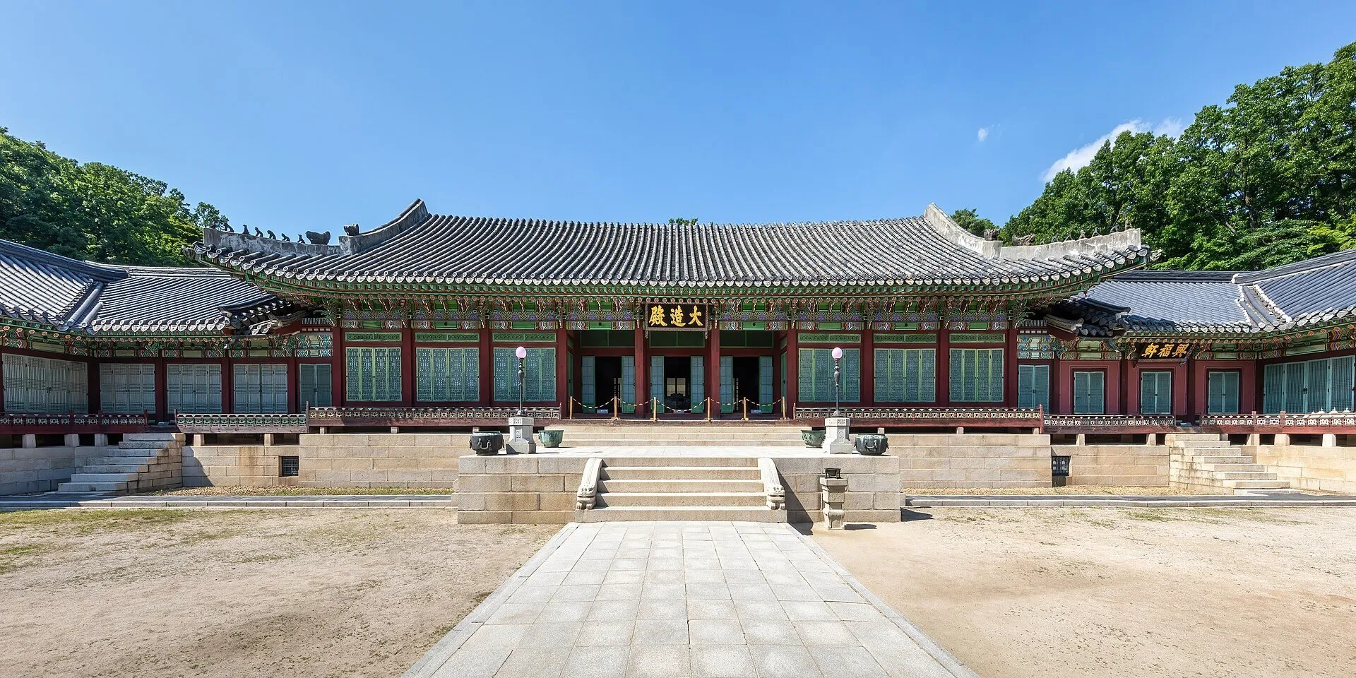 Fachada de un pabellón de Changdeokgung con tejado curvo, patio y árboles al fondo.