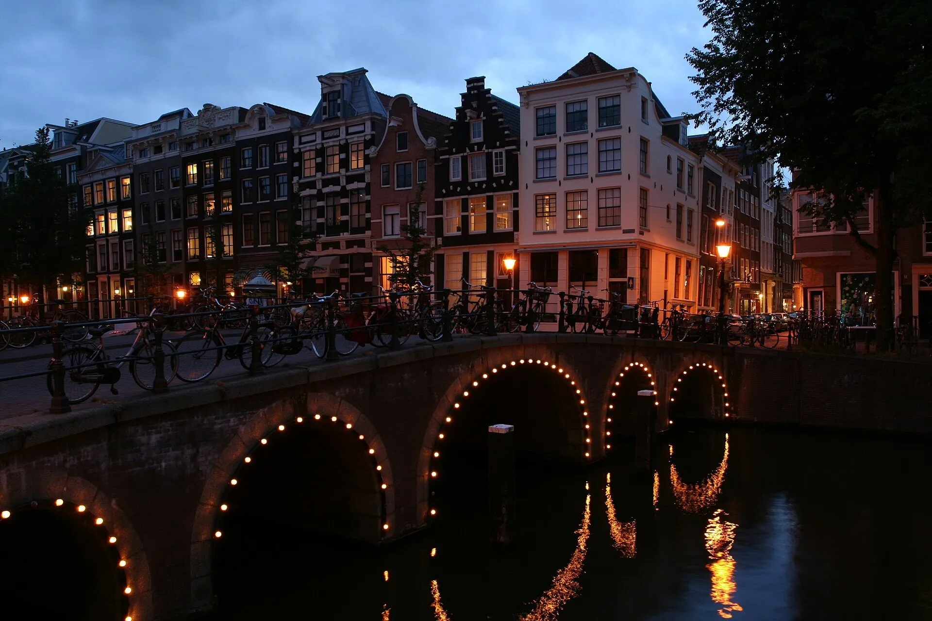 Ponte de arcos iluminados no Herengracht, com bicicletas, casas e reflexos ao anoitecer.