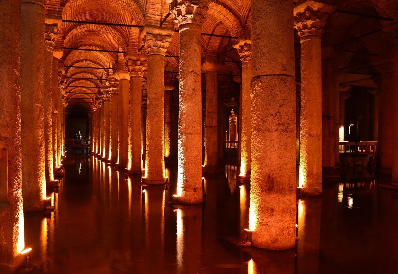 Pasillo de columnas antiguas con capiteles iluminados en naranja, bóvedas de ladrillo y reflejo en el agua de la cisterna.