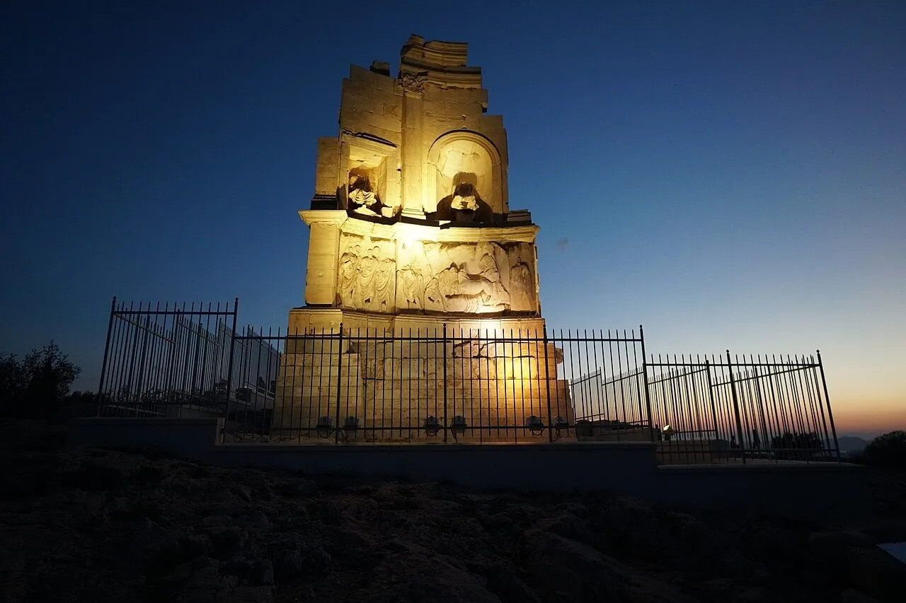 Filopappos Monument lit at dusk behind a protective fence, under a dark blue sky in Athens.