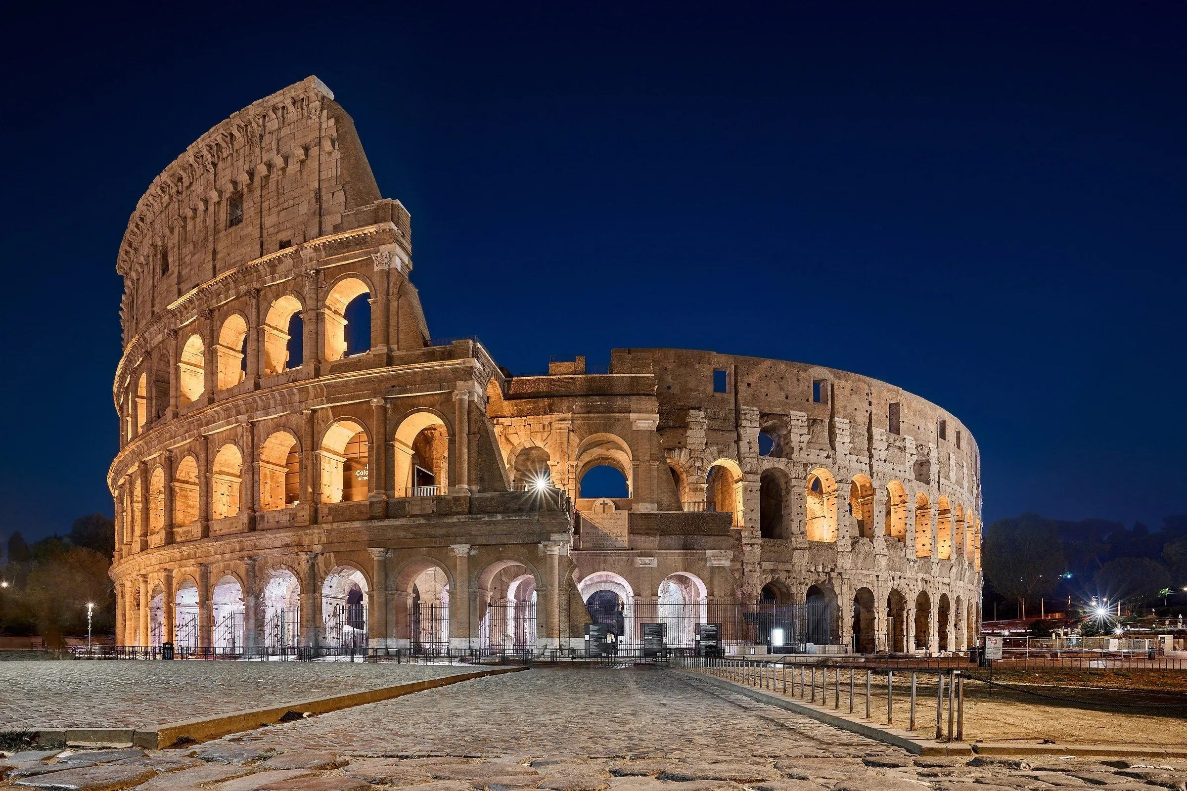 Exterior of the Colosseum in Rome lit up at dusk under a deep blue sky, with an empty square in front.