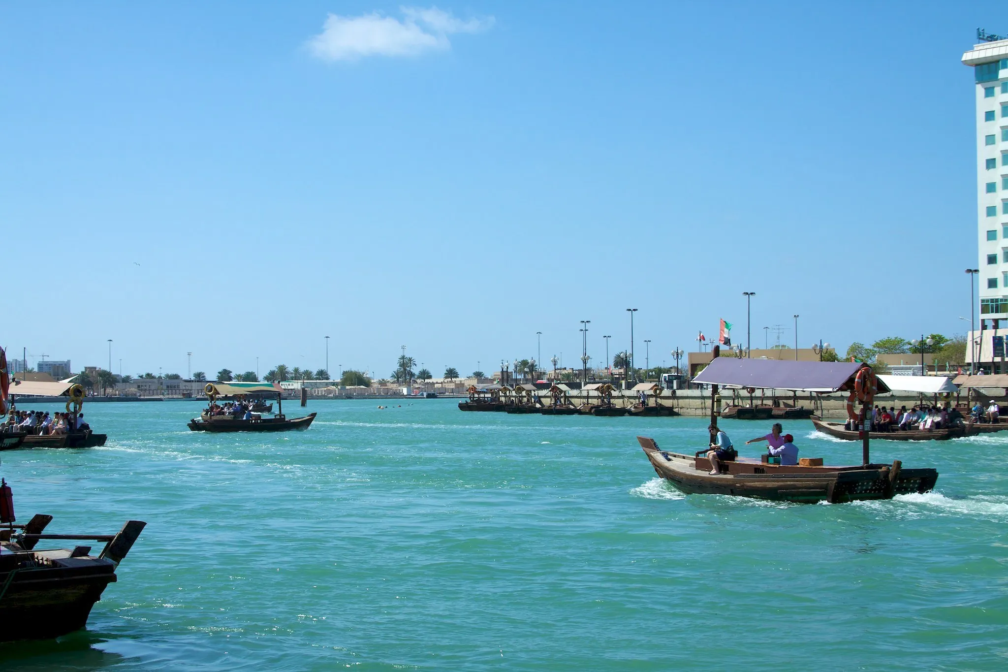 Wooden abras with passengers crossing the turquoise waters of Dubai Creek under a blue sky, with the Deira shore behind.