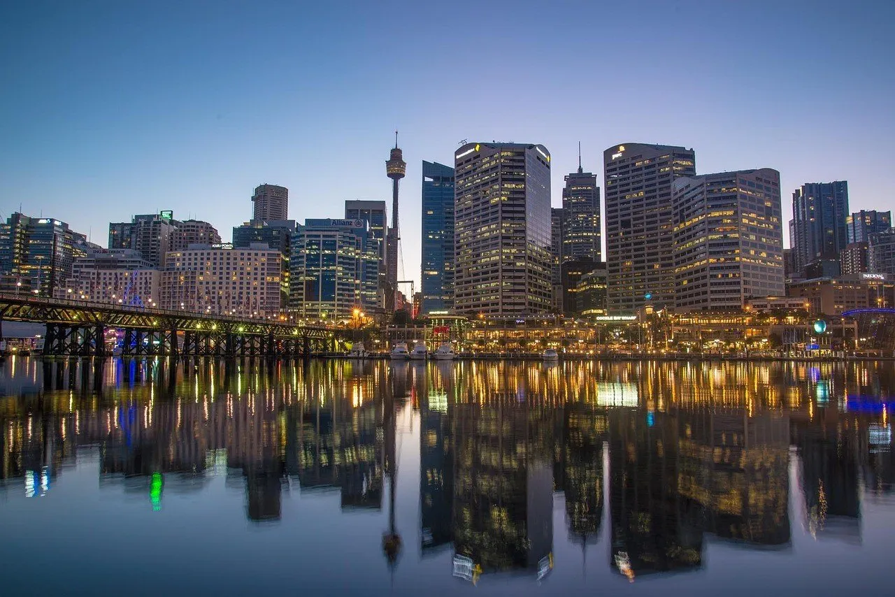 Illuminated buildings and a pedestrian bridge reflected in the calm waters of Darling Harbour under a blue dusk sky.
