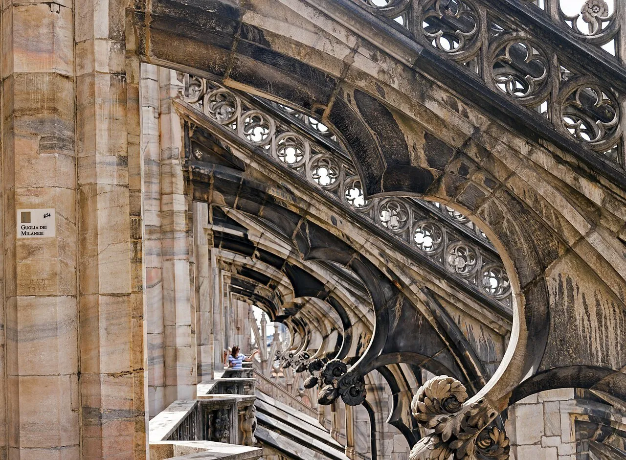 Marble flying buttresses and balustrades on the terraces of the Milan Duomo, with visitors in the background.