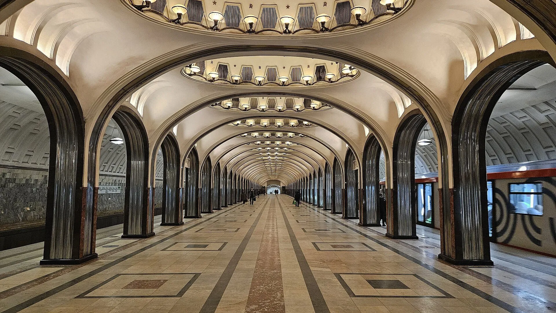 Interior de la estación Mayakovskaya de Moscú, con arcos, nichos iluminados en el techo y un tren a la derecha.