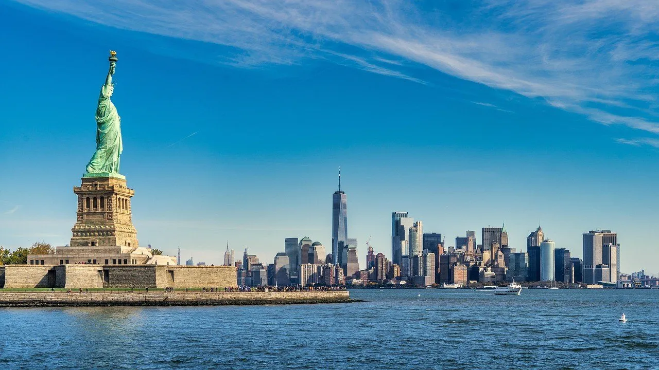Estátua da Liberdade sobre o pedestal na Liberty Island, com o horizonte de Lower Manhattan ao fundo sob céu azul.
