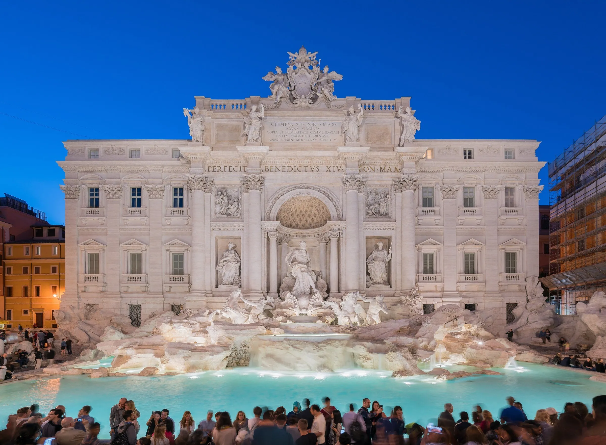 Facade of the Trevi Fountain lit up at dusk, with its turquoise basin of water and visitors at the edge.