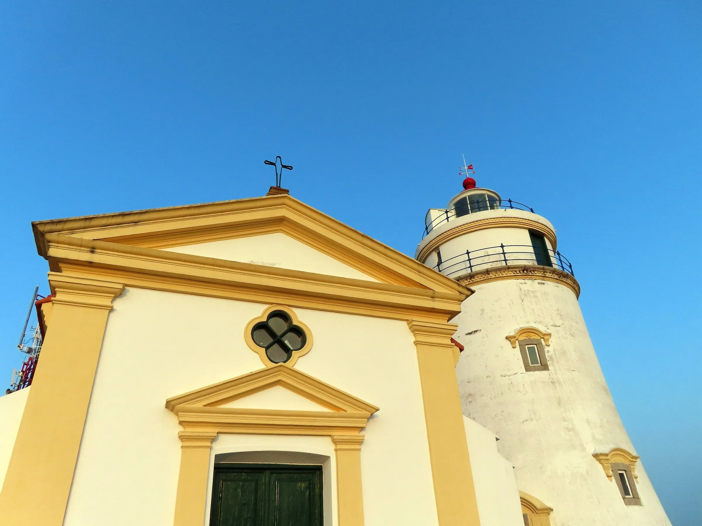 Capilla de fachada blanca y amarilla junto al Faro de Guia, torre blanca con reja azul, contra un cielo azul despejado.