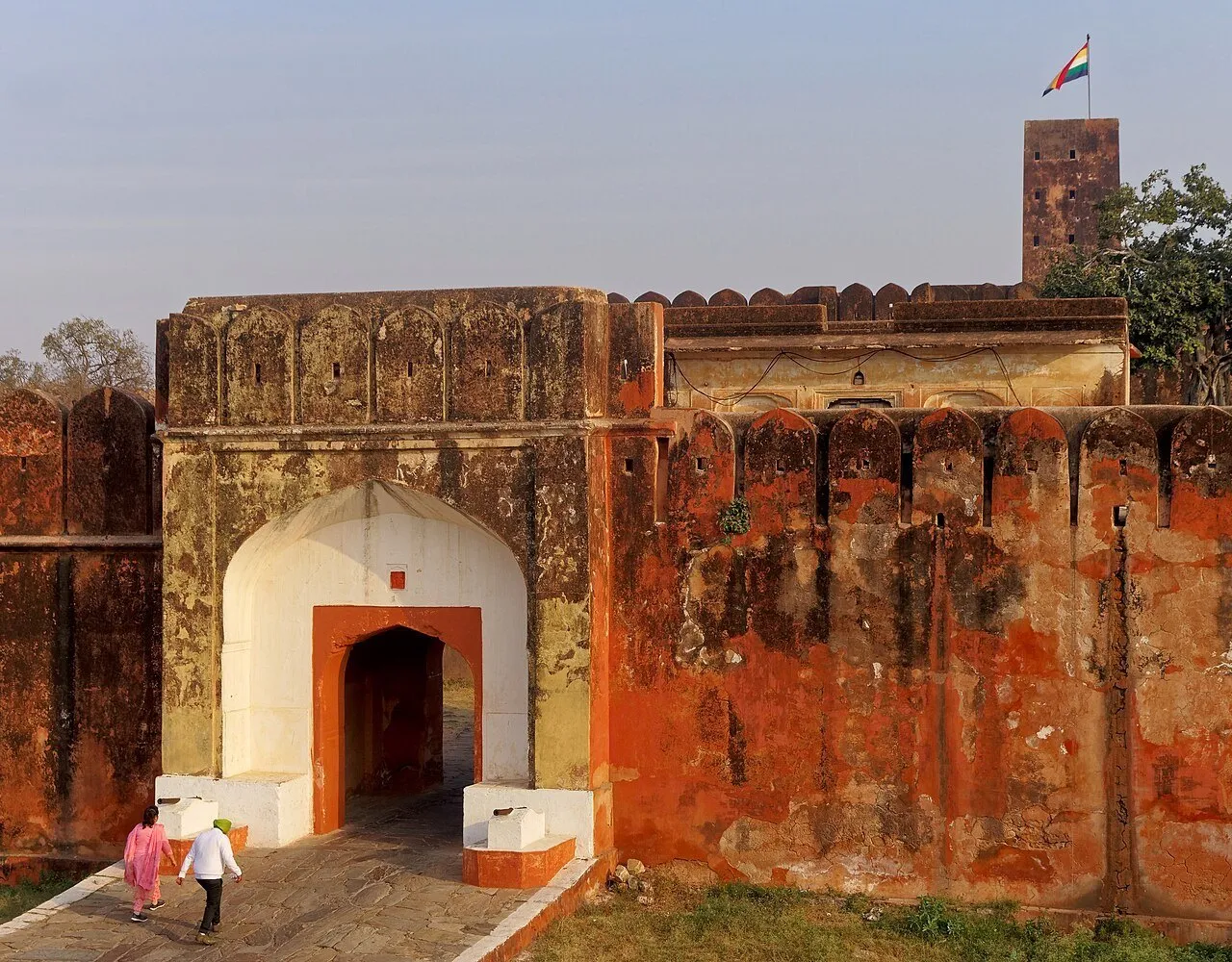 Portal claro nas muralhas avermelhadas de Jaigarh, com dois visitantes e uma torre com bandeira ao fundo.