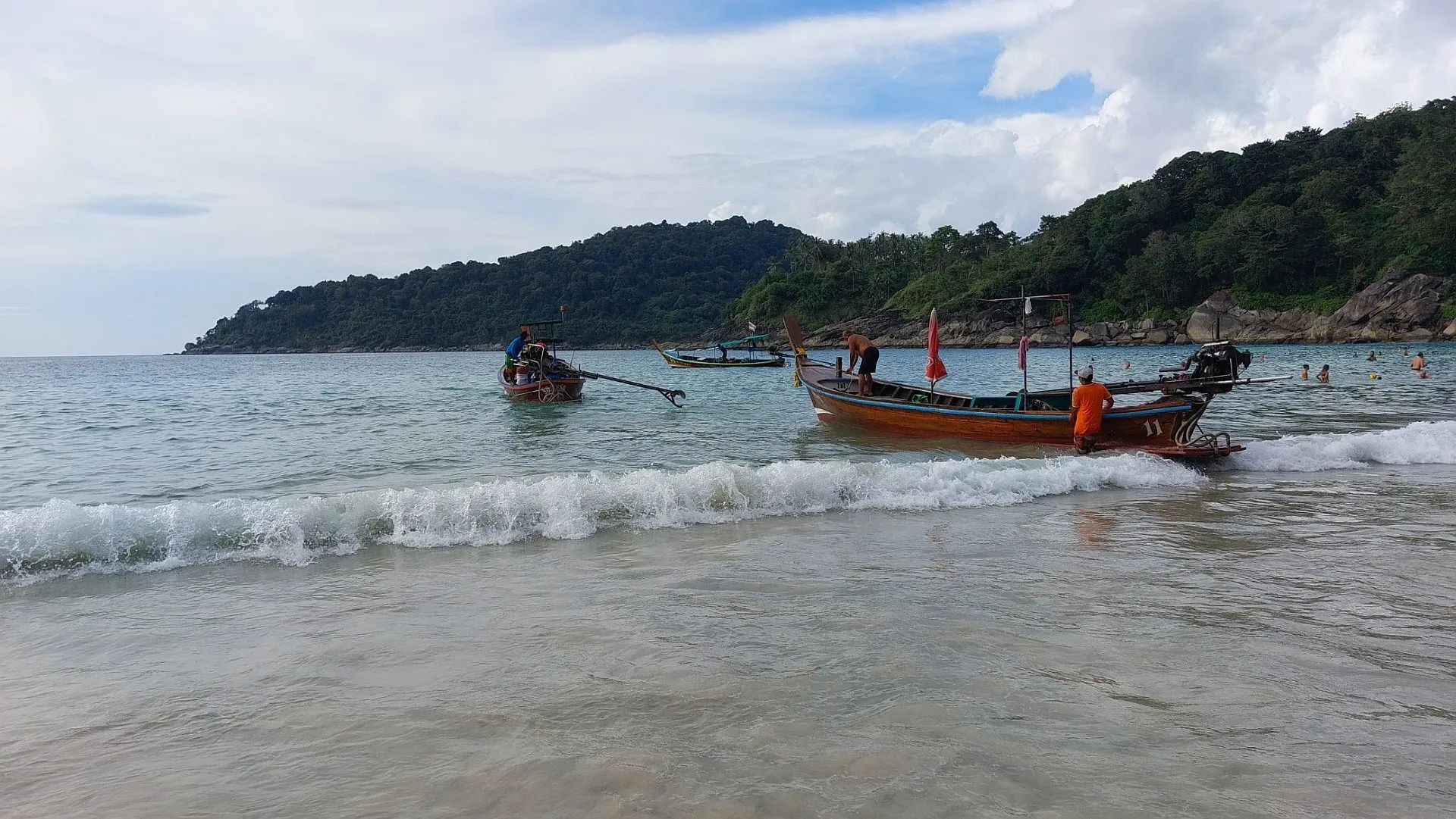Barcos long-tail na arrebentação da Freedom Beach, em Phuket, com colina coberta de mata e céu com nuvens ao fundo.