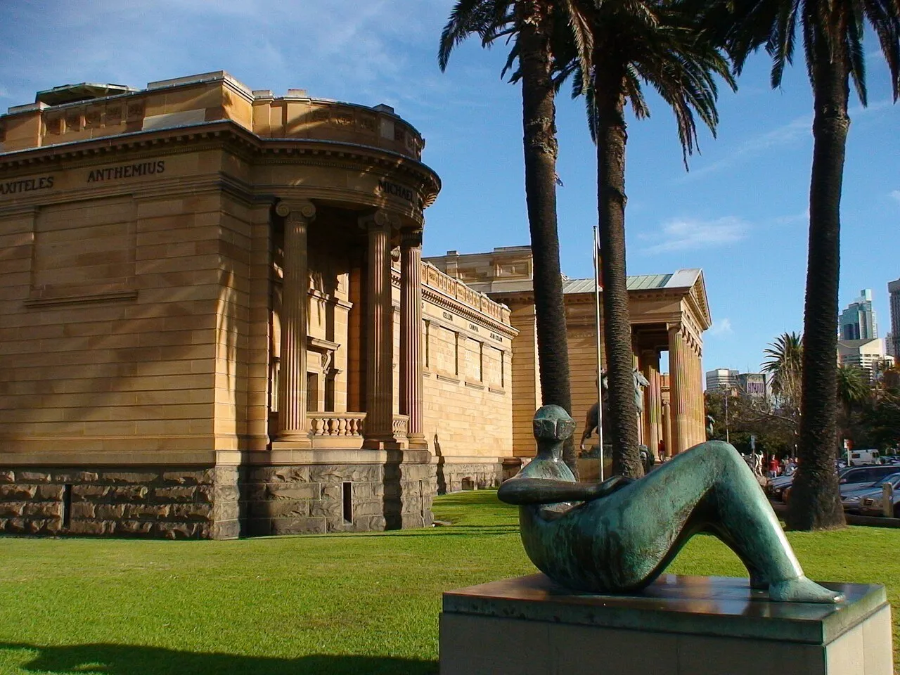 Stone exterior of the Art Gallery of New South Wales, with columns, palm trees and a reclining sculpture.