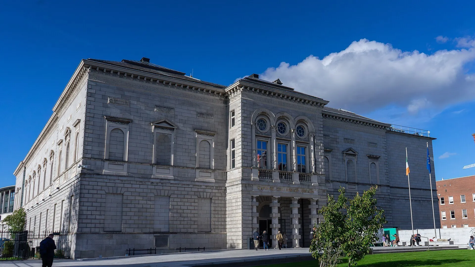 Fachada de piedra de la Galería Nacional de Irlanda con ventanas circulares, banderas y gente ante la entrada.