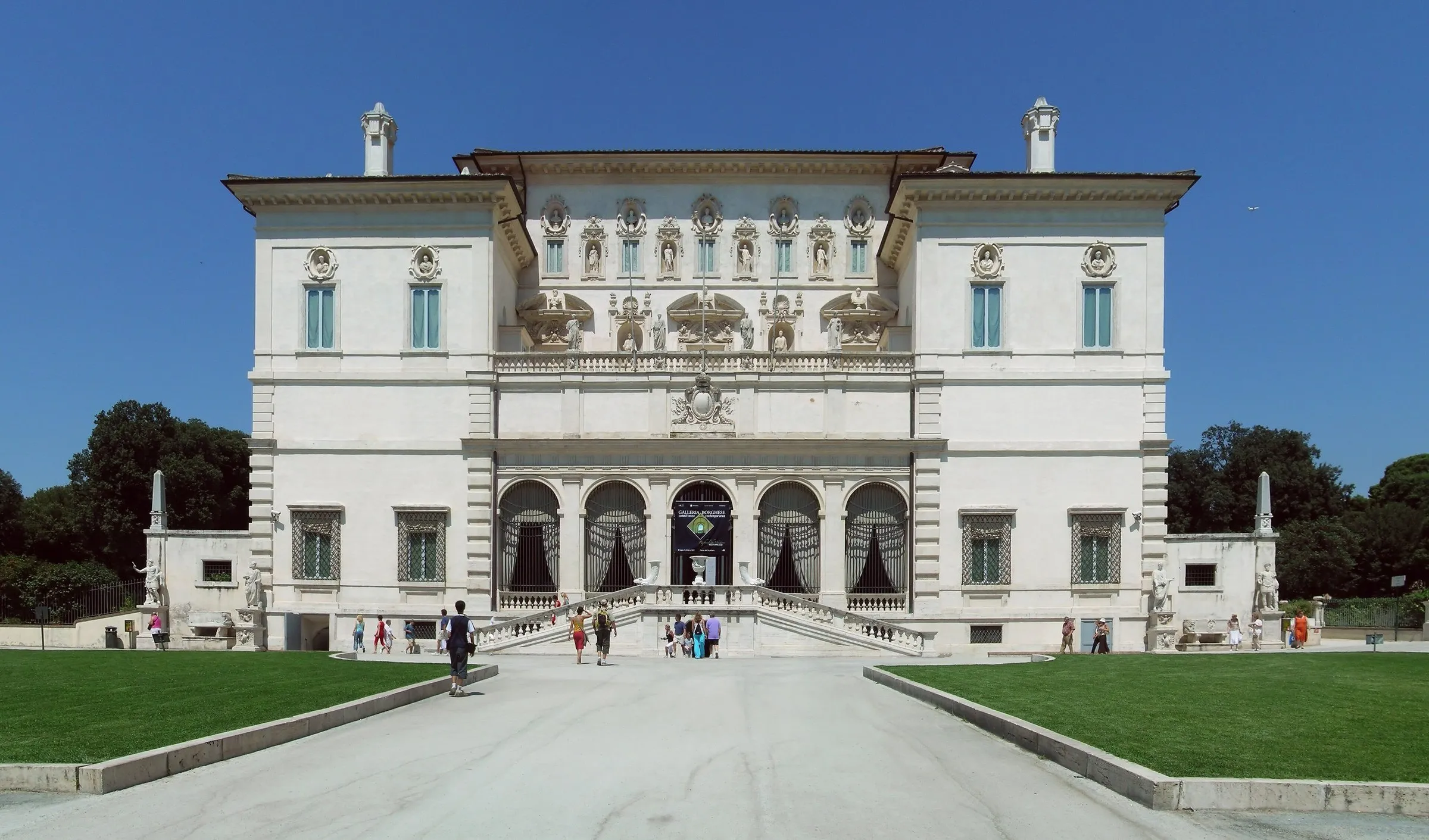 White facade of the Galleria Borghese, with an entrance staircase and lawn in the foreground under a blue sky.