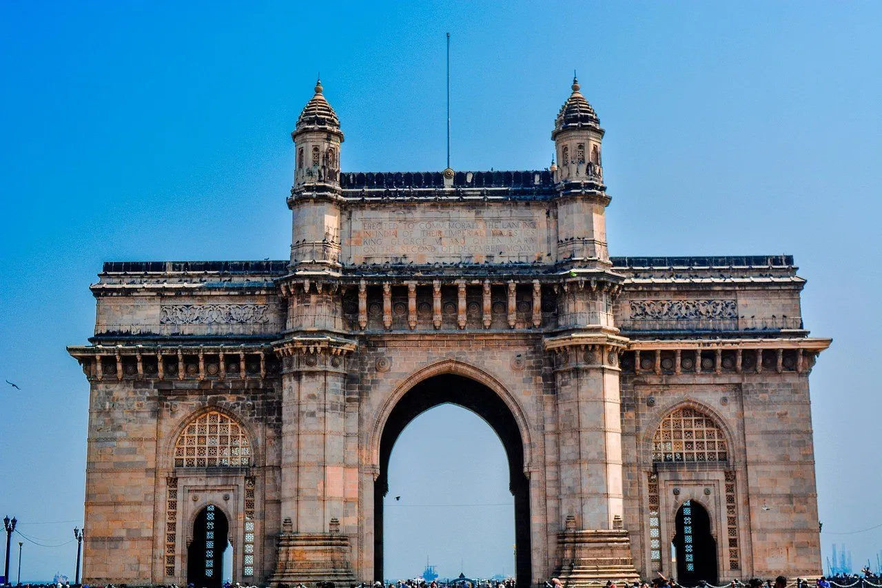 Fachada frontal do Gateway of India, em Mumbai, arco de pedra amarelada com torres laterais, contra céu azul.