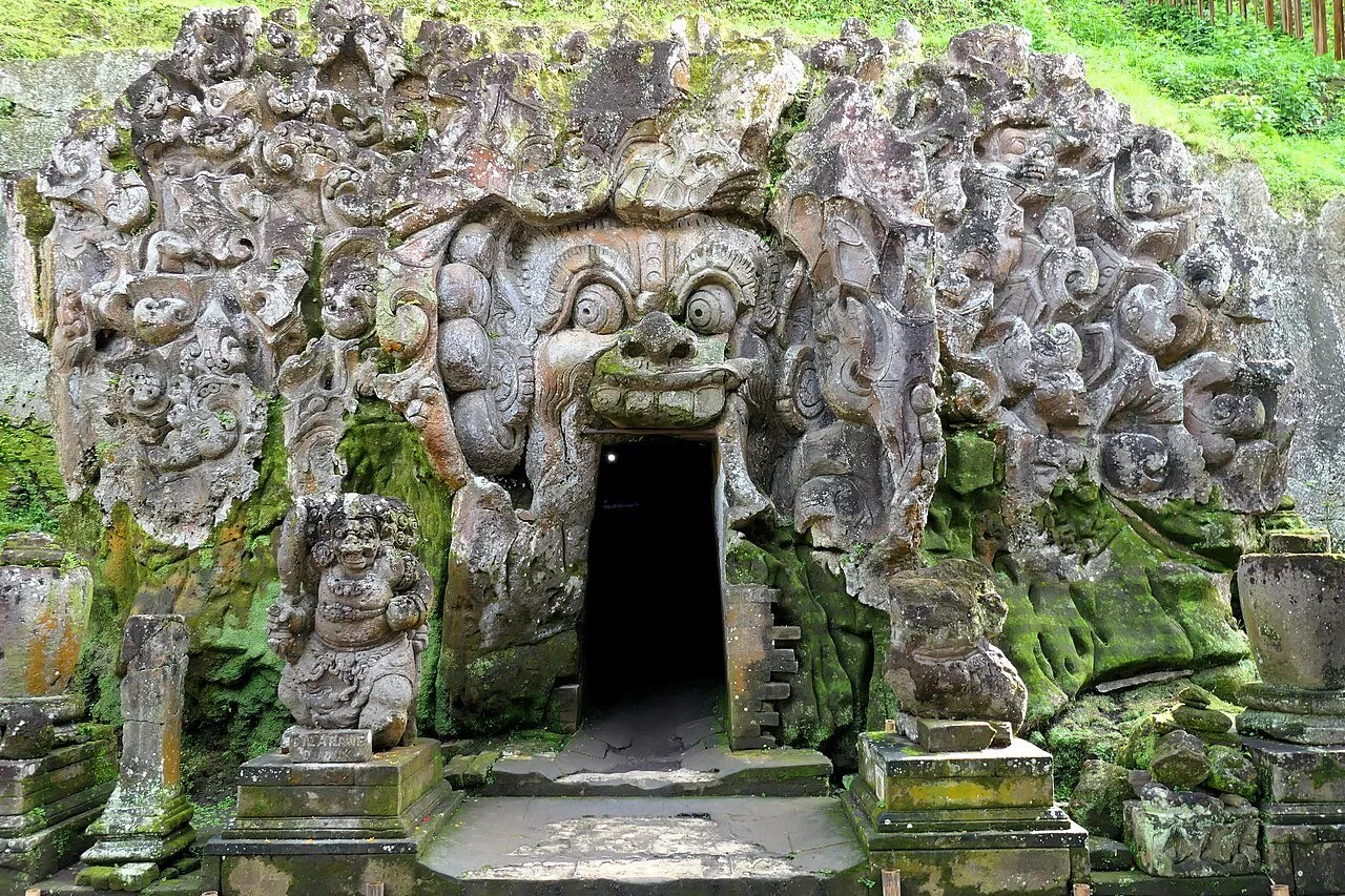 Entrada oscura de la cueva de Goa Gajah enmarcada por un rostro y relieves esculpidos en la piedra, con musgo.