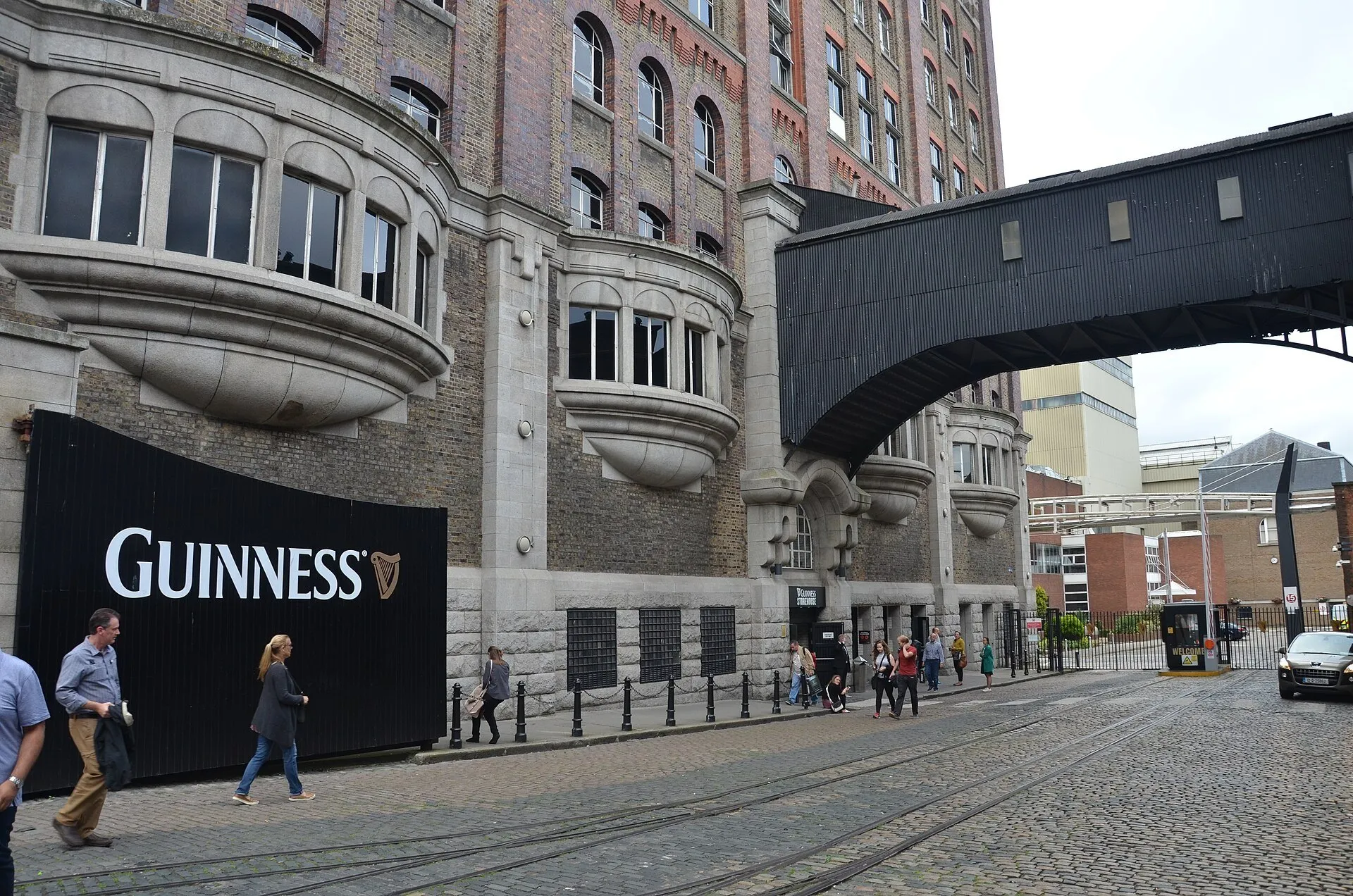 Fachada del Guinness Storehouse con ventanas curvas, pasarela elevada, visitantes y calle empedrada.