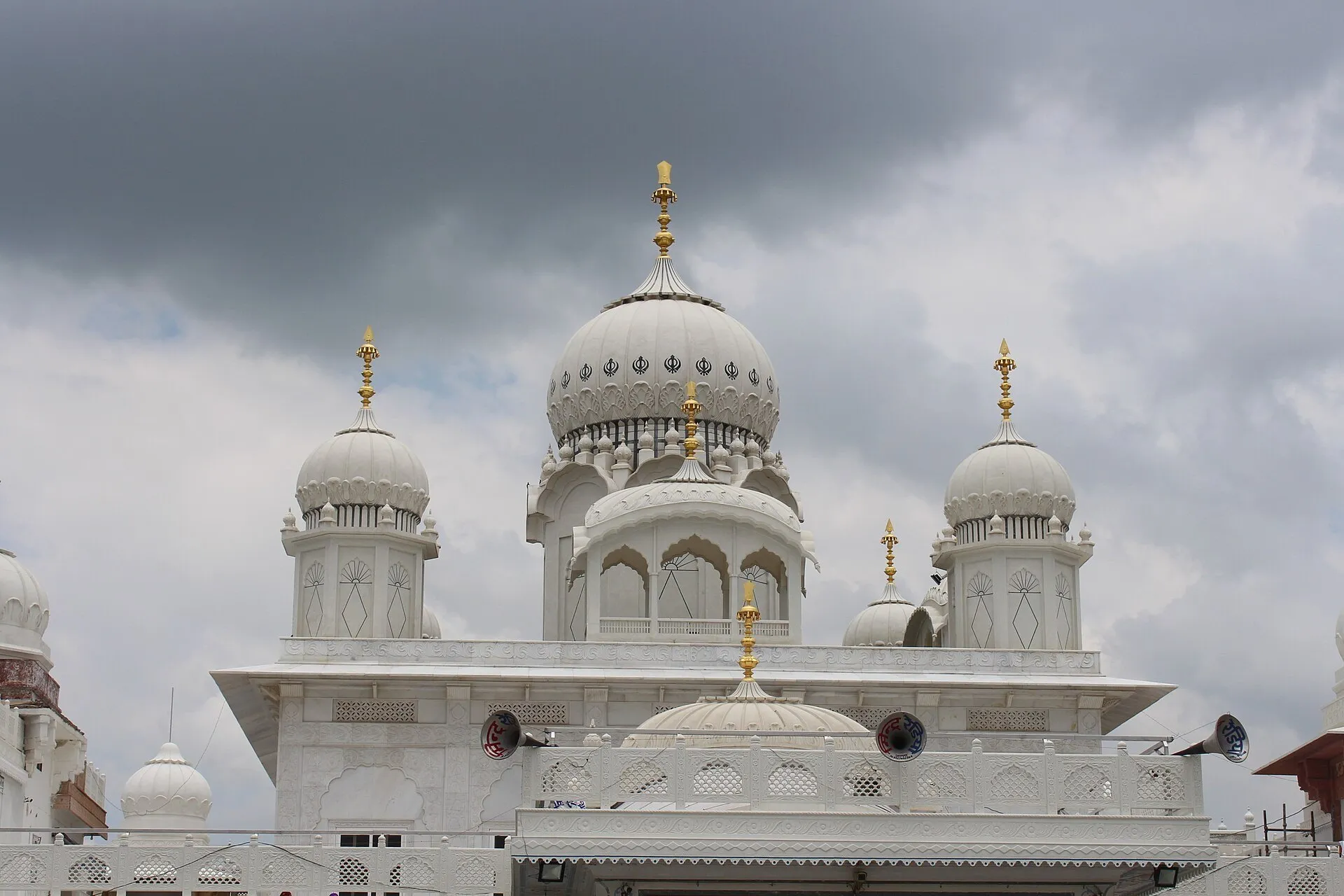 Cúpulas blancas, remates dorados y fachada trabajada del gurdwara Guru ka Tal, bajo nubes.
