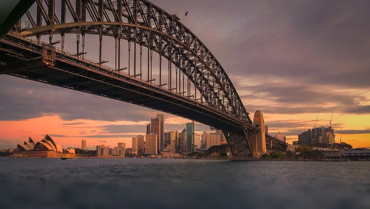 Harbour Bridge structure over the harbor, with the Opera House on the left and buildings behind under a late-afternoon sky.