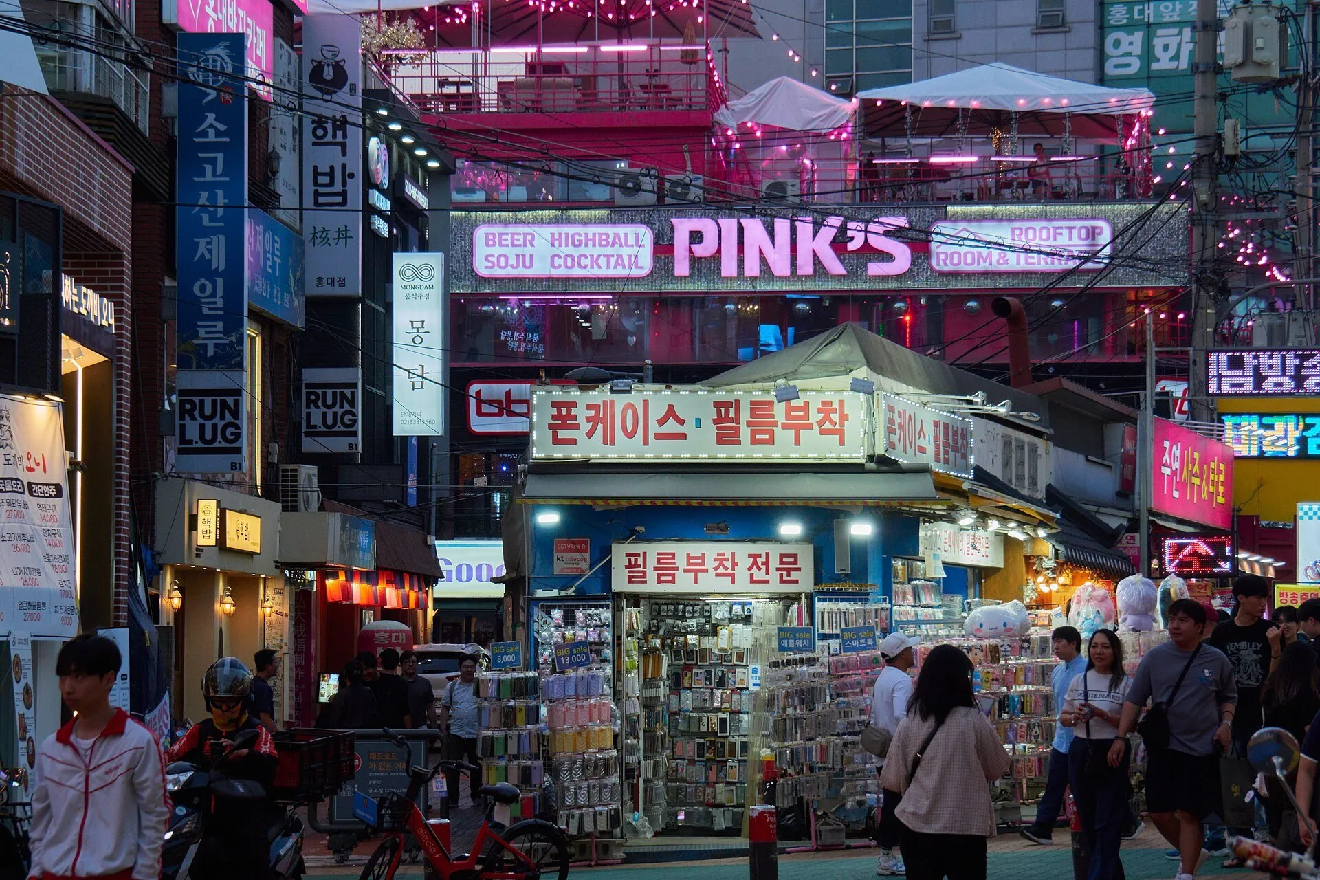 Tiendas y letreros iluminados en una calle de Hongdae, con peatones y un puesto de accesorios.