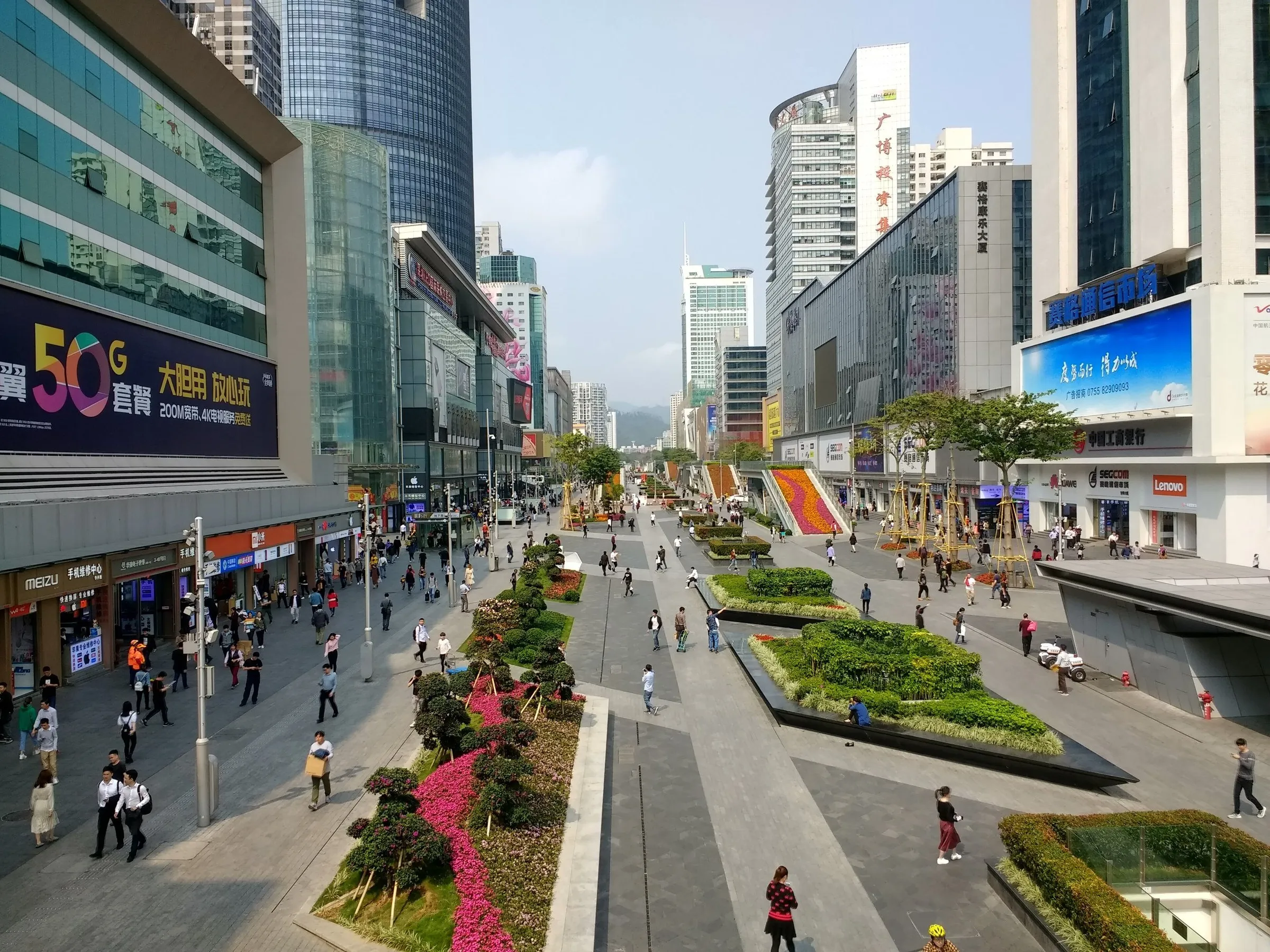 Rua de pedestres de Huaqiangbei vista de uma passarela, com canteiros floridos e prédios de lojas de eletrônicos.