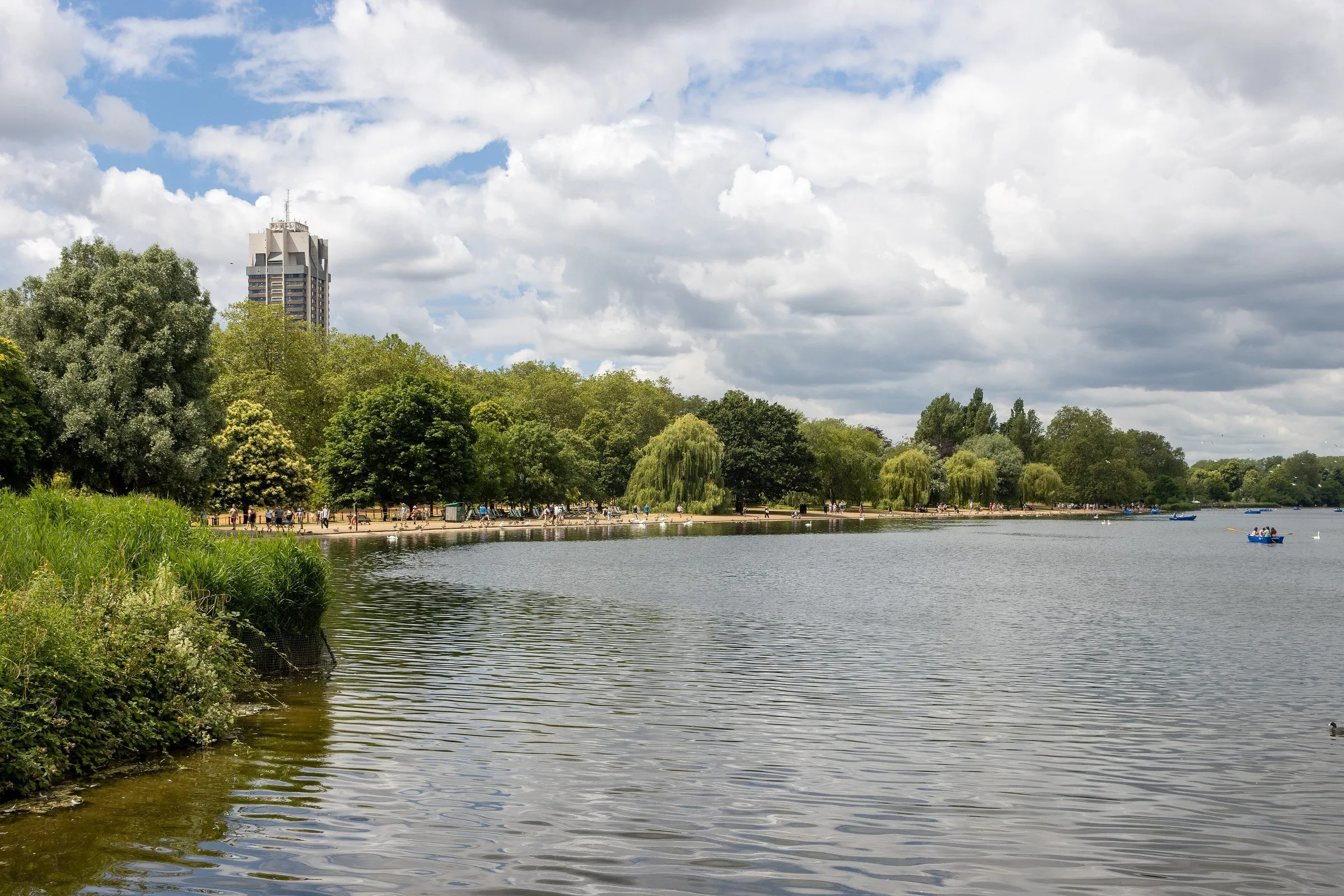 Serpentine lake in Hyde Park, with trees along the shore, people on the bank and rowboats in the distance under cloudy skies.