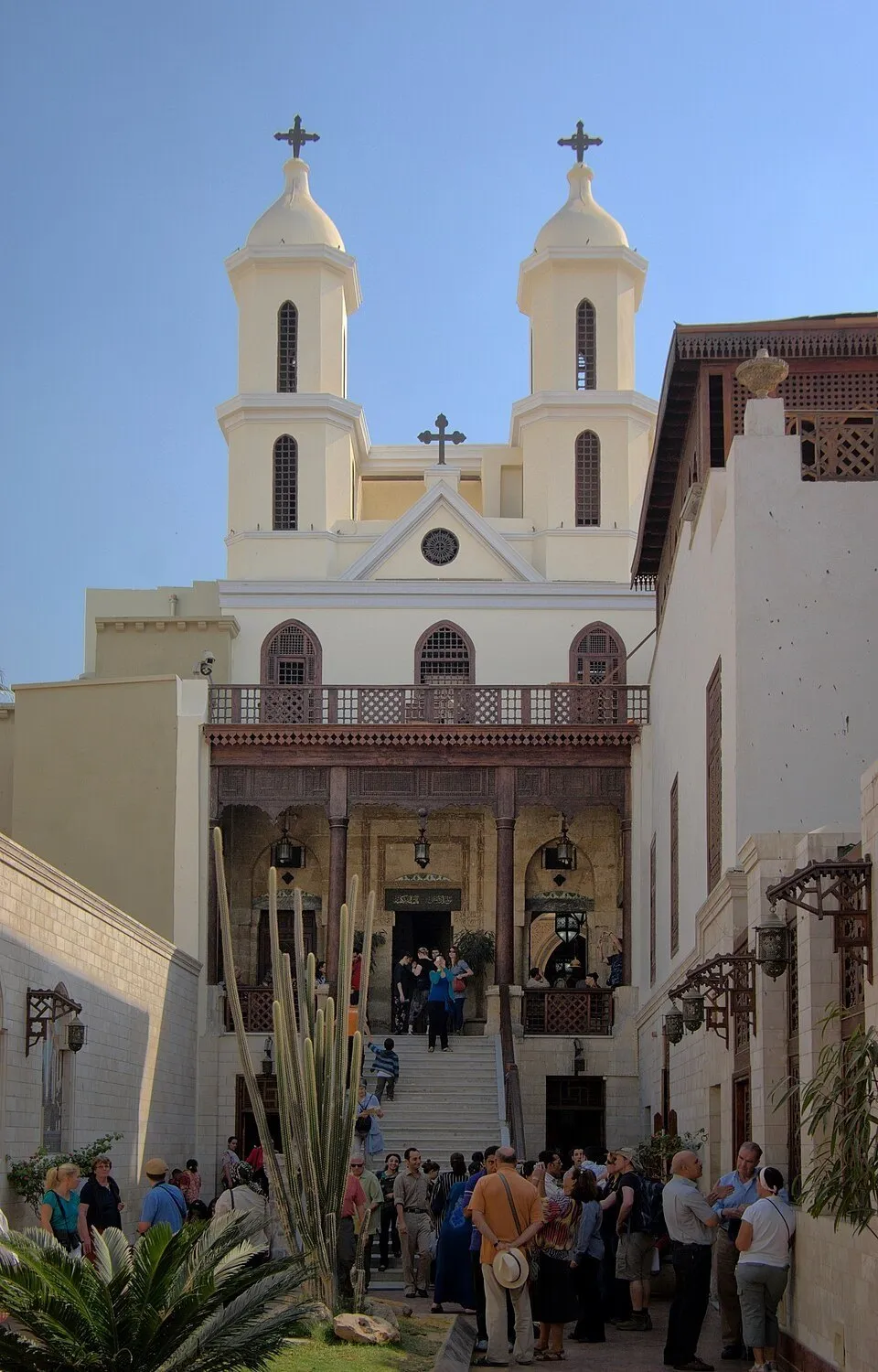 Fachada da Igreja Suspensa no Cairo, com duas torres claras, cruzes, escadaria e visitantes no pátio.