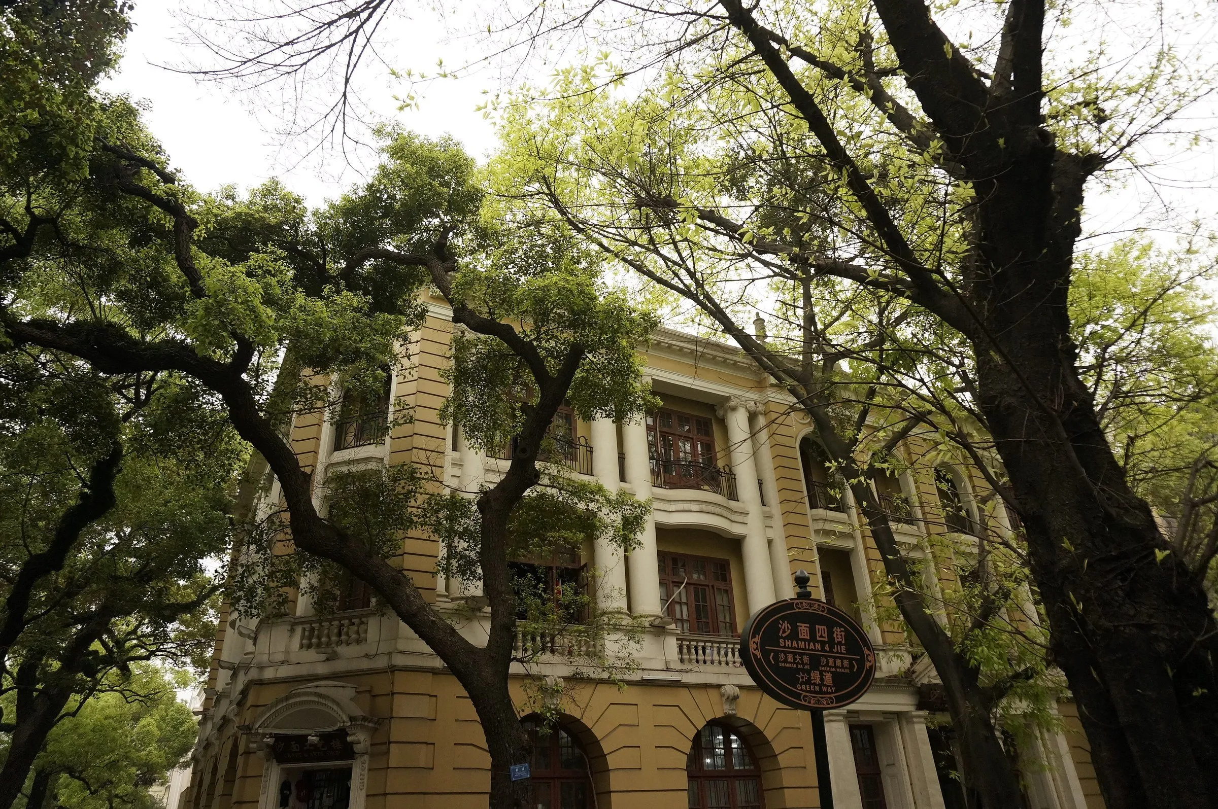 Fachada amarilla con columnas y balcones entre árboles en la isla de Shamian, con un letrero de calle en primer plano.