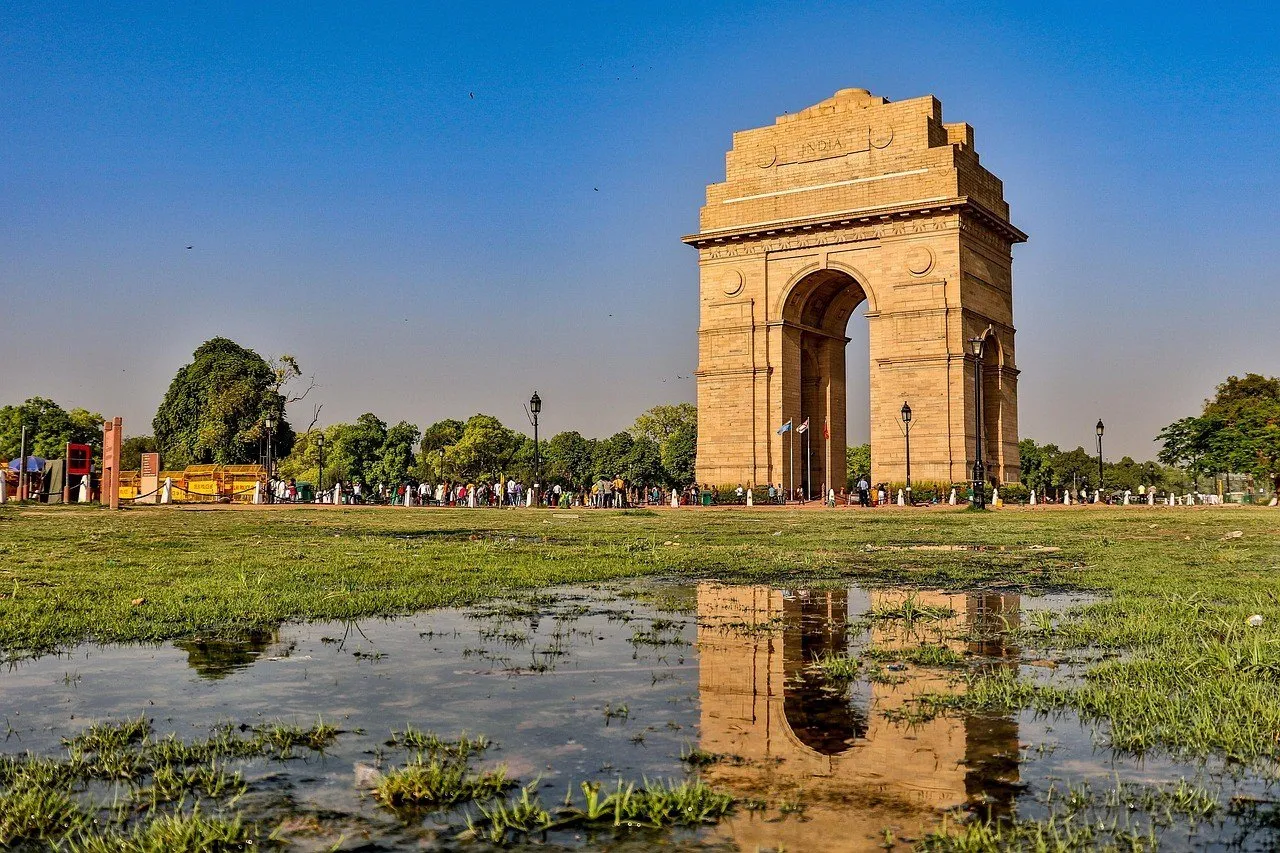 Puerta de la India, en Nueva Delhi, de arenisca clara bajo cielo azul, con su reflejo en un charco del césped.