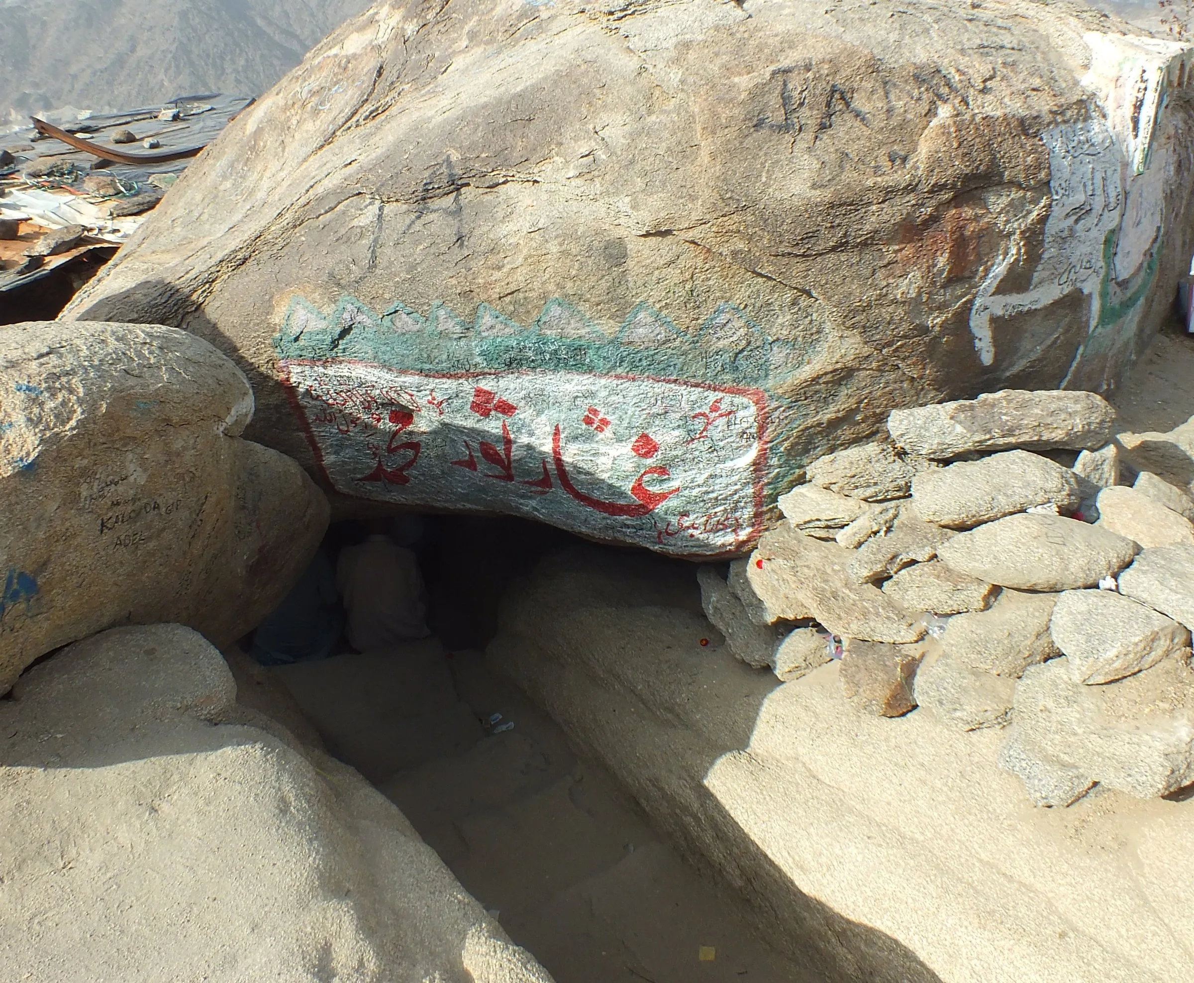 Entrada baixa da caverna de Thawr, com inscrições nas rochas e pedras ao redor, sob luz do dia.