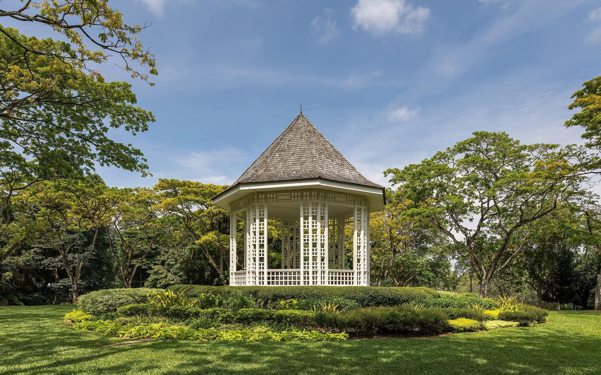 Coreto branco de madeira cercado por árvores e canteiros, sob céu azul com nuvens, no Jardim Botânico de Singapura.